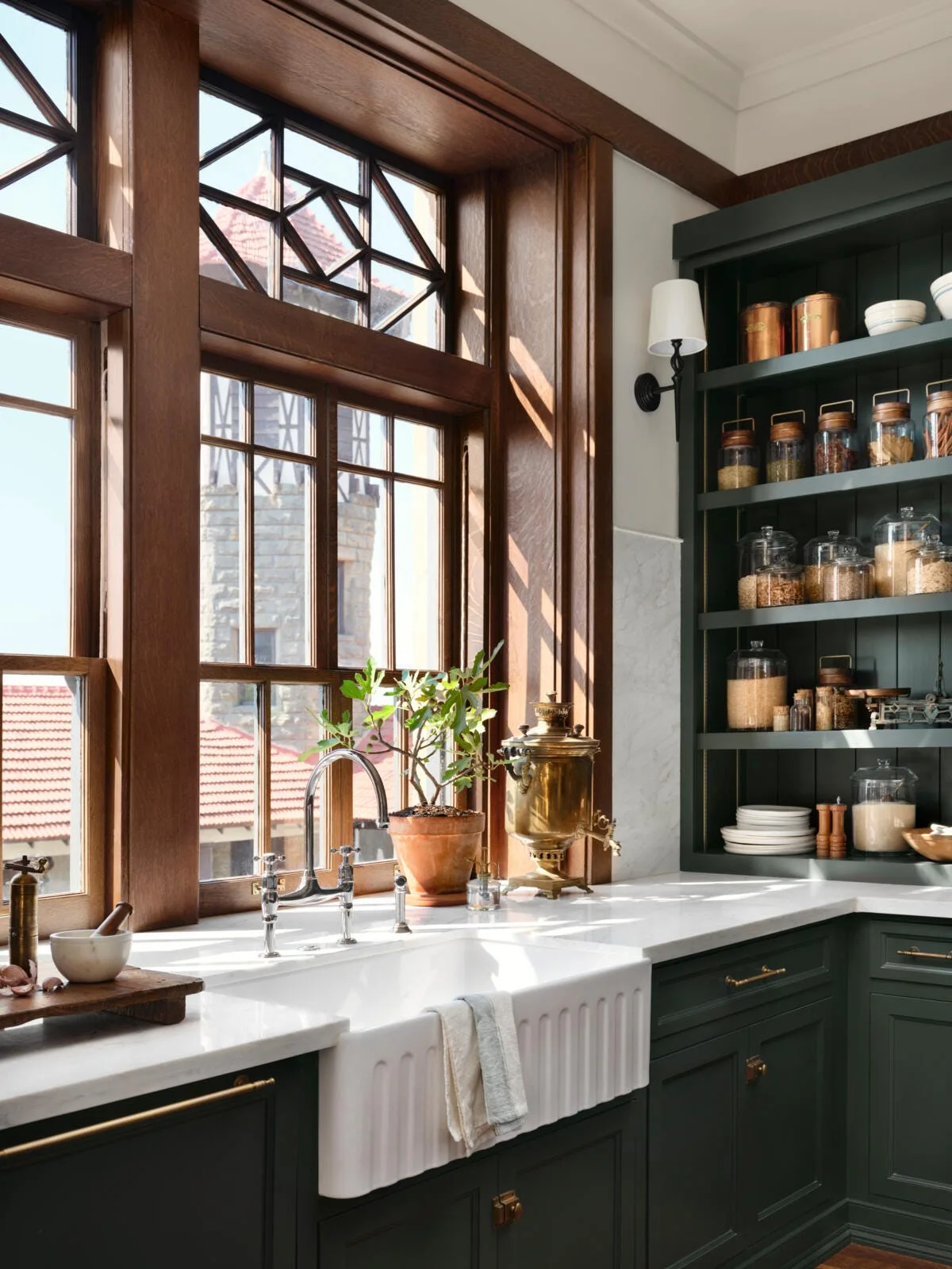 A kitchen features a white fluted farmhouse sink, dark green cabinetry, open shelving filled with glass jars, and a stained wood window.