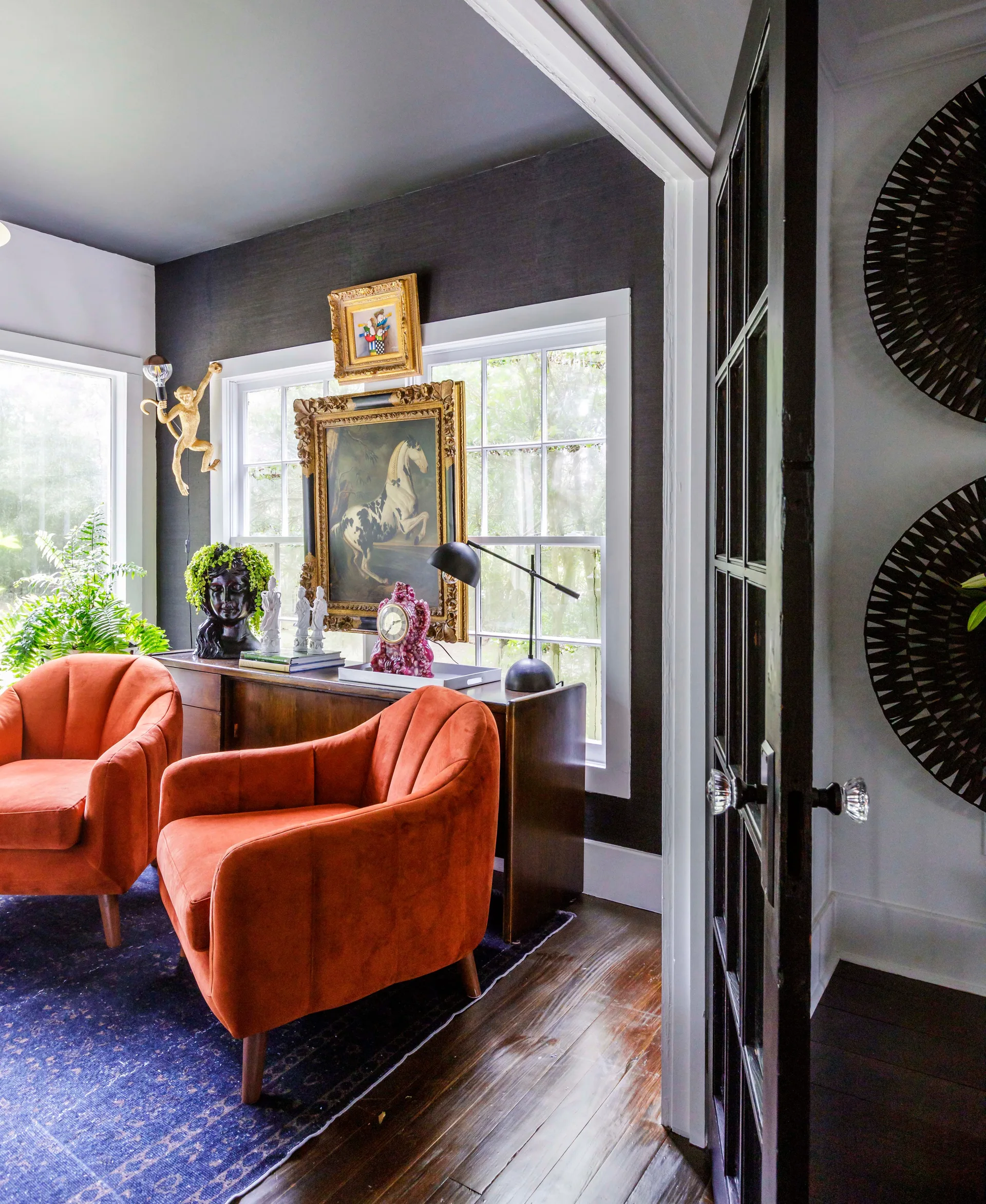 Two orange velvet armchairs sit on a blue patterned rug in an eclectic room with dark charcoal walls, multi-pane windows, and a wood console table.