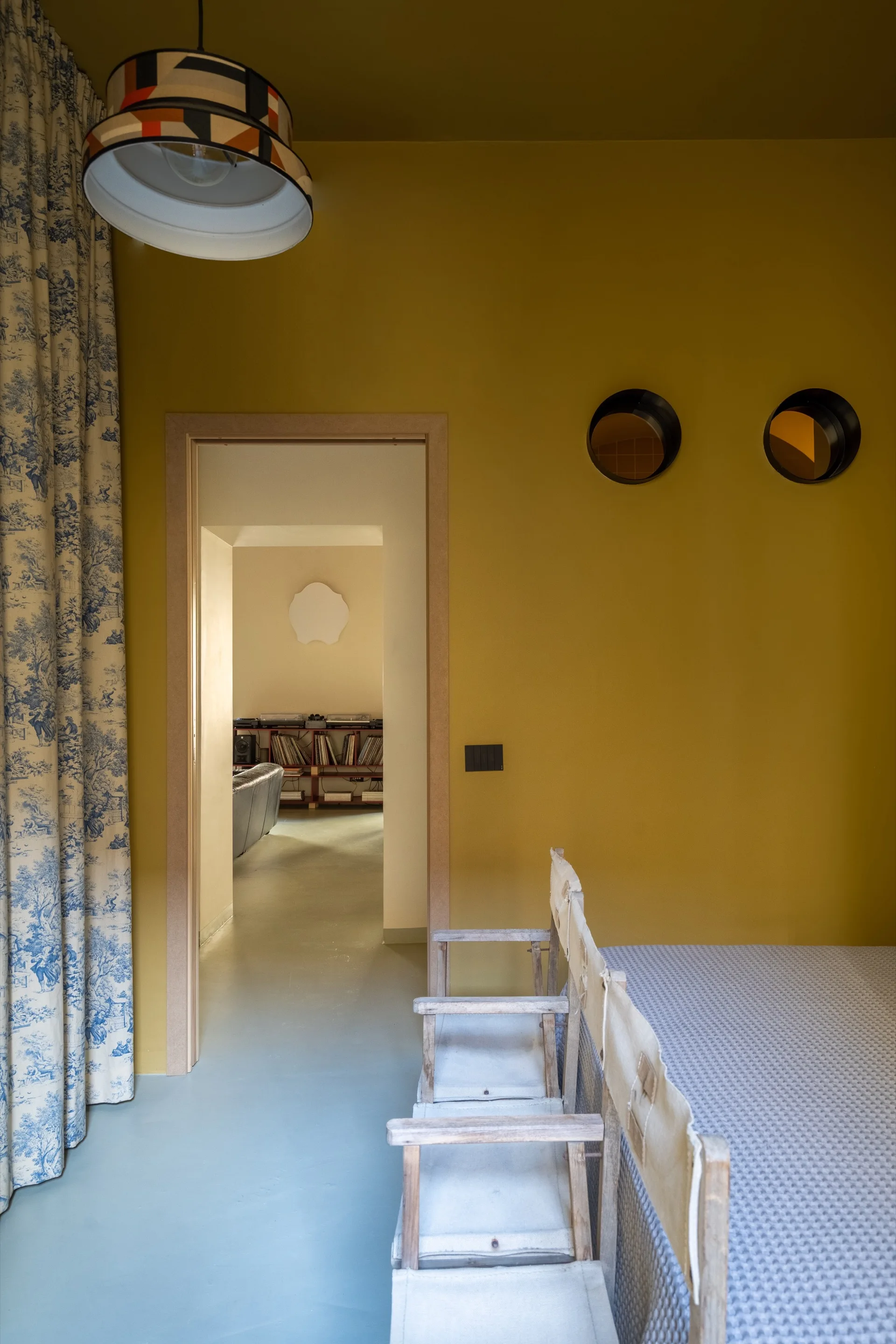 An ochre yellow room features blue and white toile curtains, director-style canvas chairs, circular porthole cutouts, and a doorway to an adjacent room.