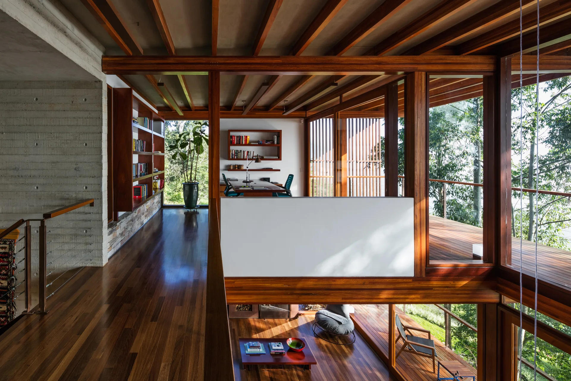 A multi-level home interior features exposed dark wood beams, a concrete wall, built-in wooden bookshelves, and floor-to-ceiling windows showing trees.