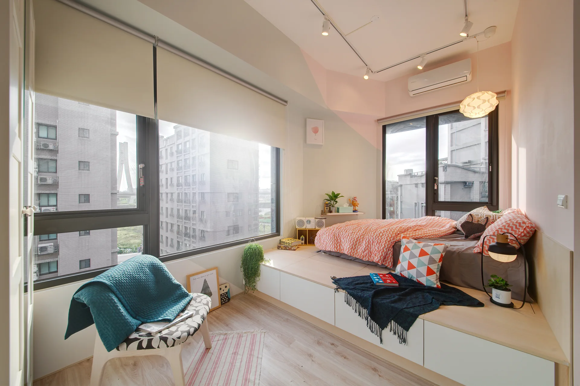 A small bedroom featuring a custom raised wooden platform bed with integrated storage drawers next to large windows and a patterned armchair.