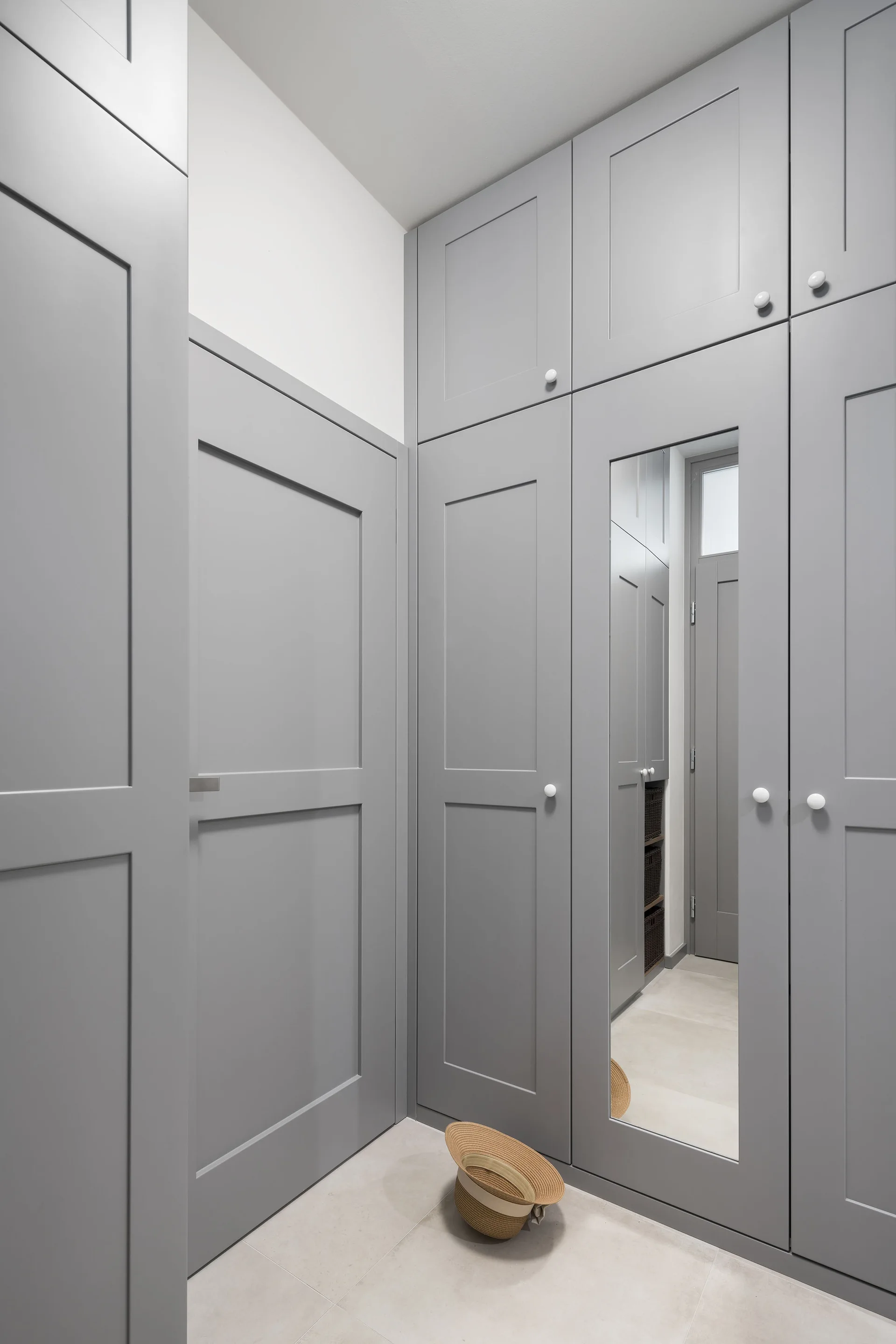 Floor-to-ceiling grey shaker cabinets with small white knobs stand in an entryway, with one door featuring an integrated full-length mirror.