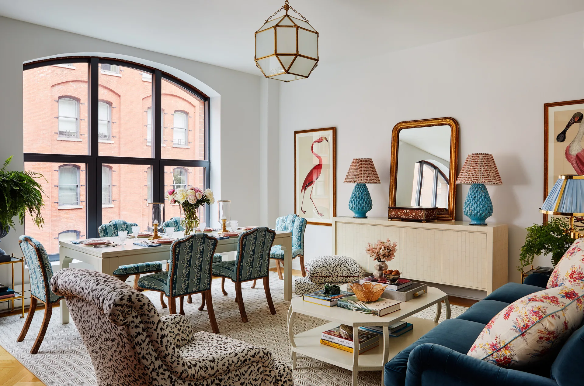 An open living and dining room features blue patterned chairs, a velvet sofa, and a large black-framed arched window.