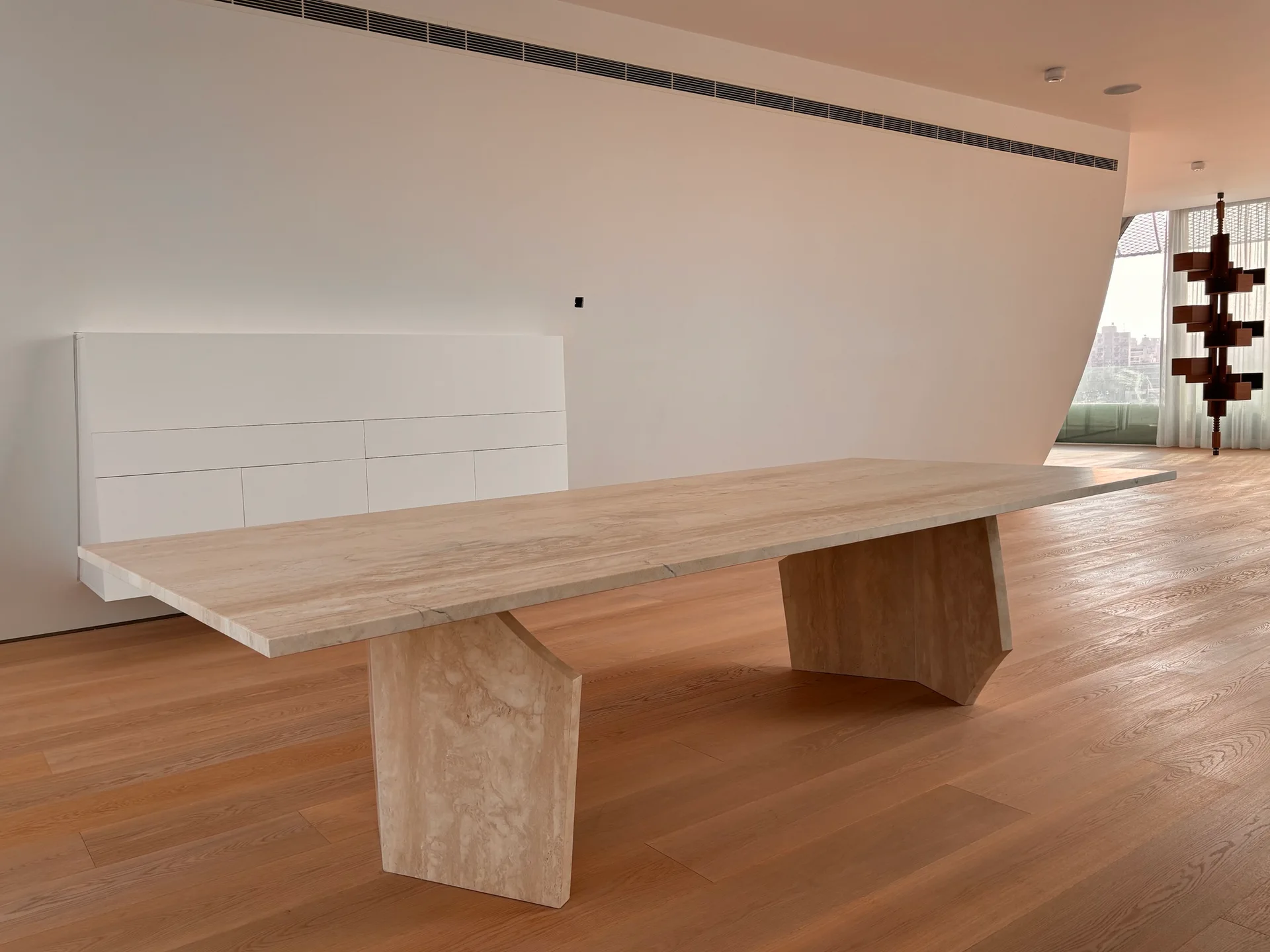 A large travertine stone dining table sits on wide-plank wood floors in a minimalist room with a low-profile white cabinet on the wall.