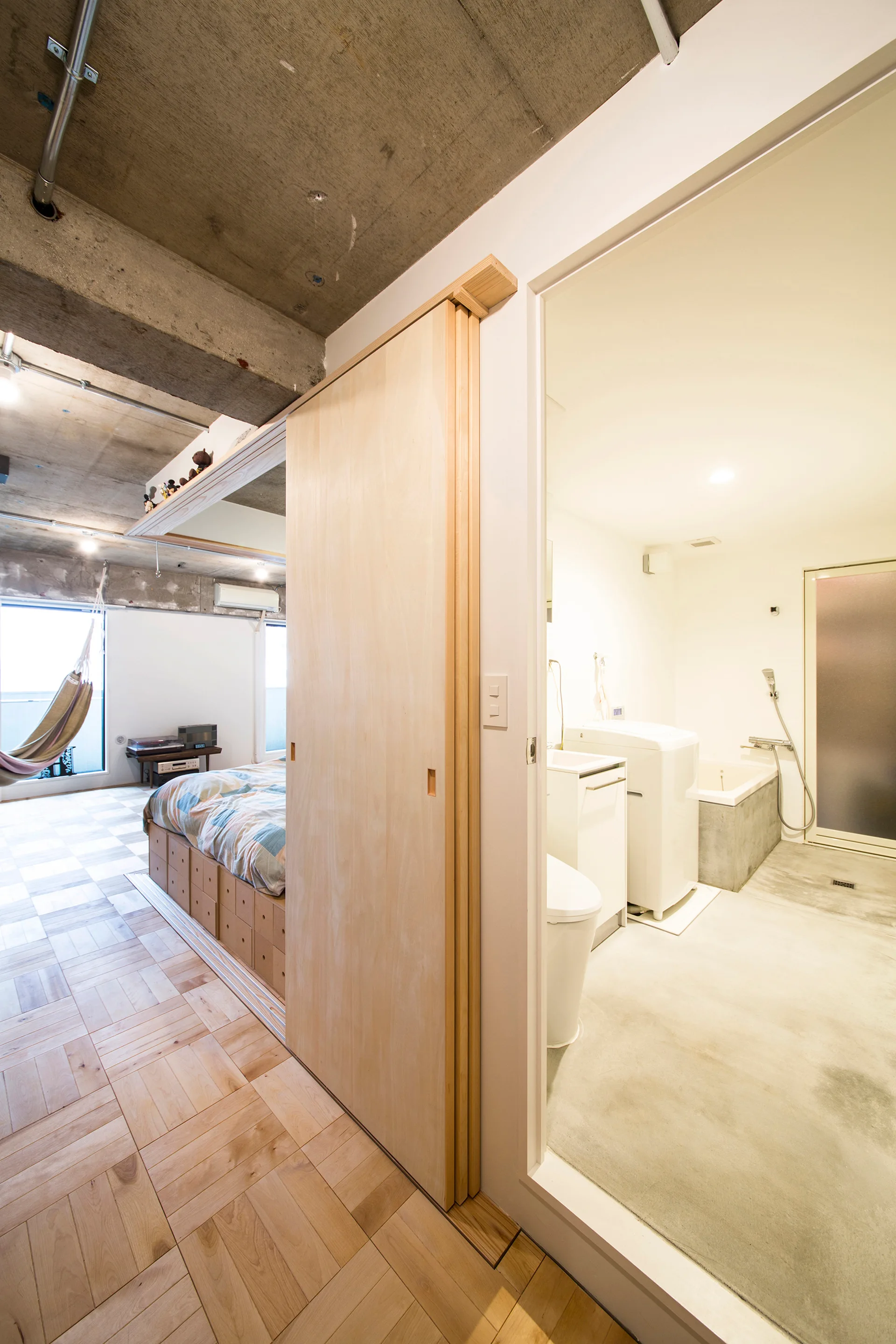 A minimalist studio with a bedroom and bathroom separated by a sliding wooden door, featuring parquet flooring and an exposed concrete ceiling.