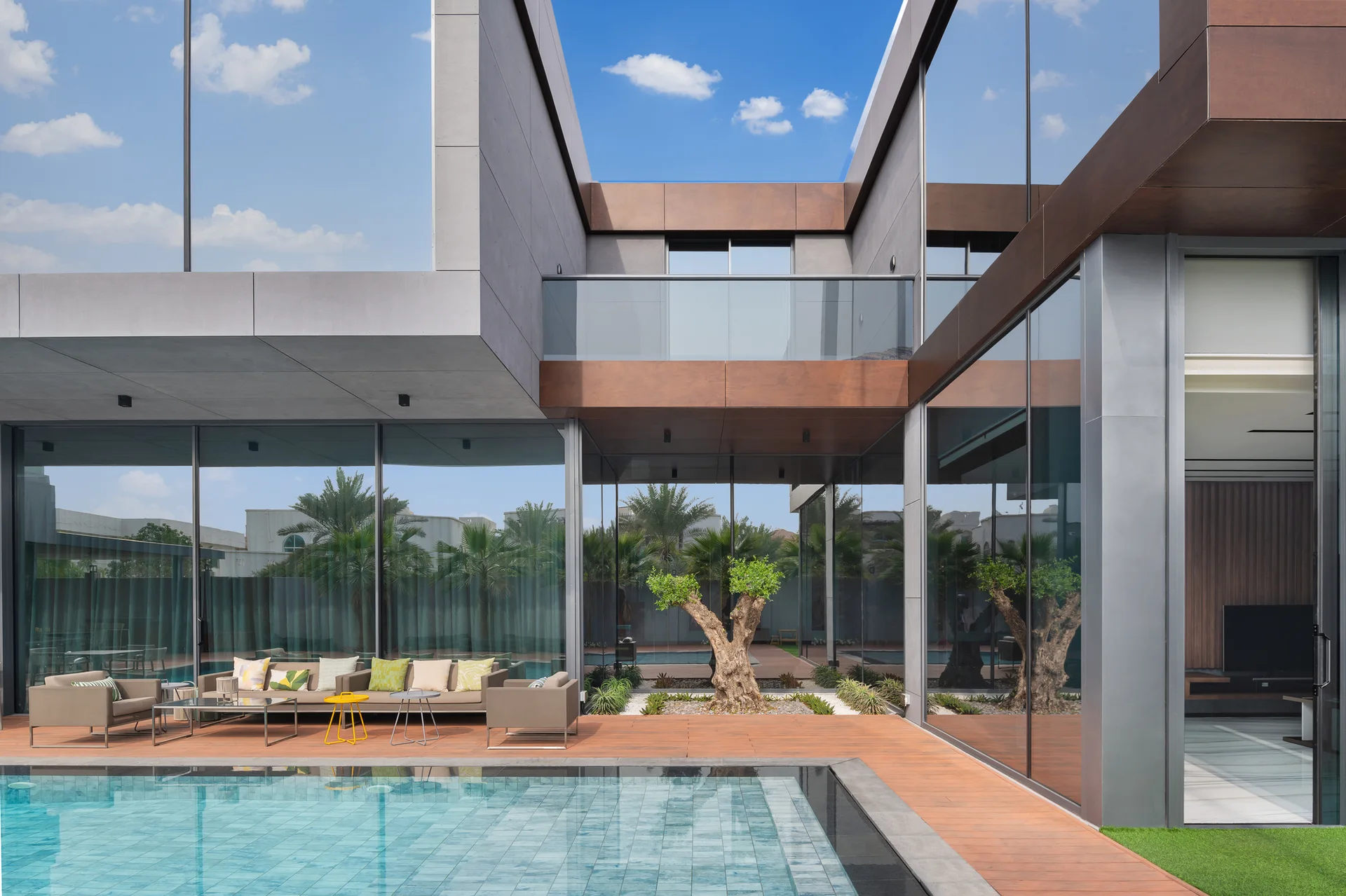 A modern poolside patio features beige lounge furniture on a wooden deck, reflective glass walls, and a central courtyard with a sculptural tree.
