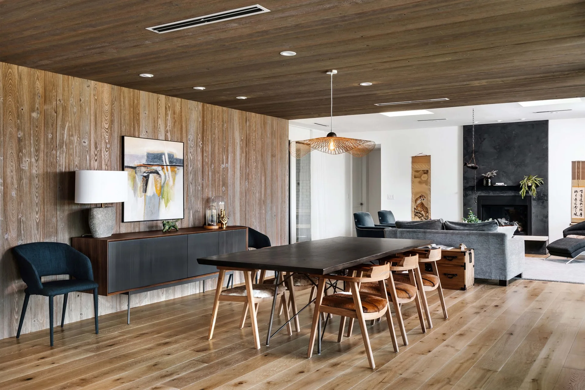 An open-concept dining room features a dark wood table with cowhide chairs, set against vertical wood wall paneling and a matching wood ceiling.