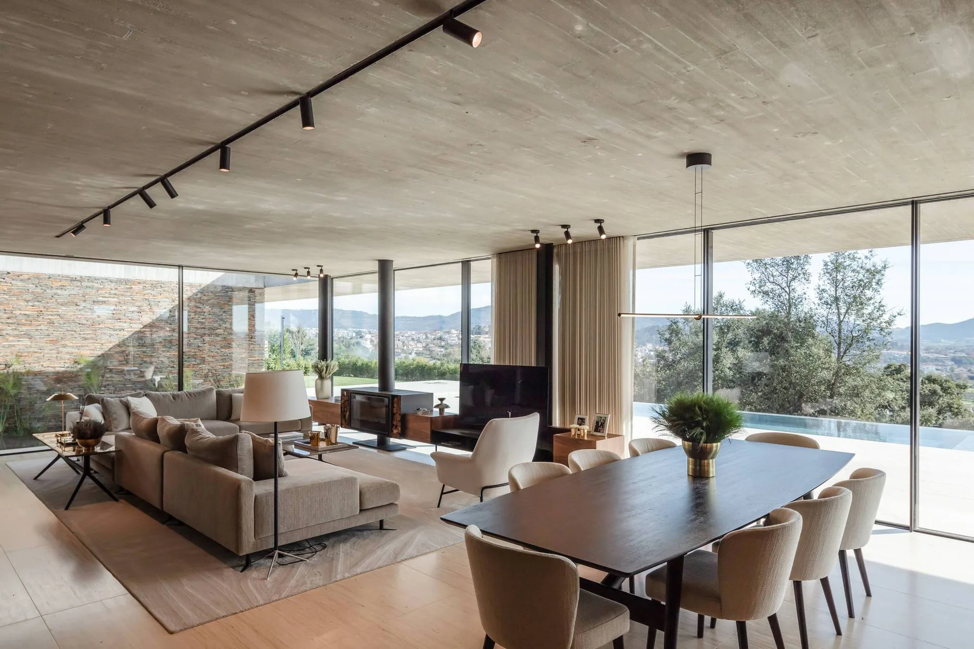 An open-plan living and dining room with an exposed concrete ceiling, a grey sectional sofa, a dark wood dining table, and floor-to-ceiling windows.