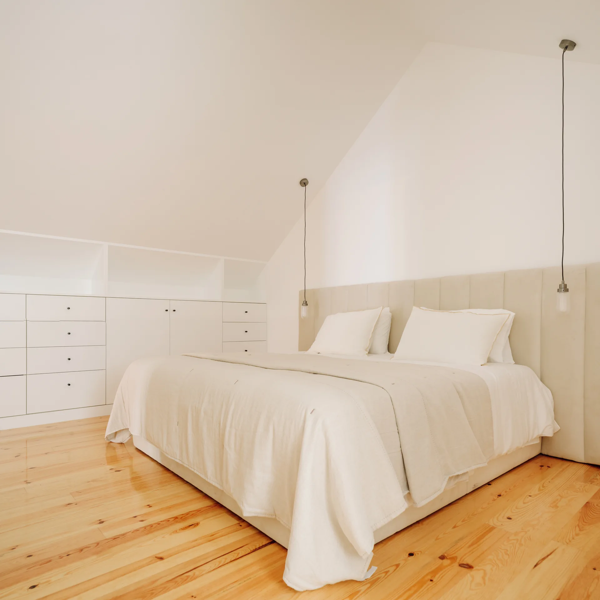 Minimalist attic bedroom with a low bed, beige headboard, white built-in drawers under a slanted ceiling, and natural wood floors.