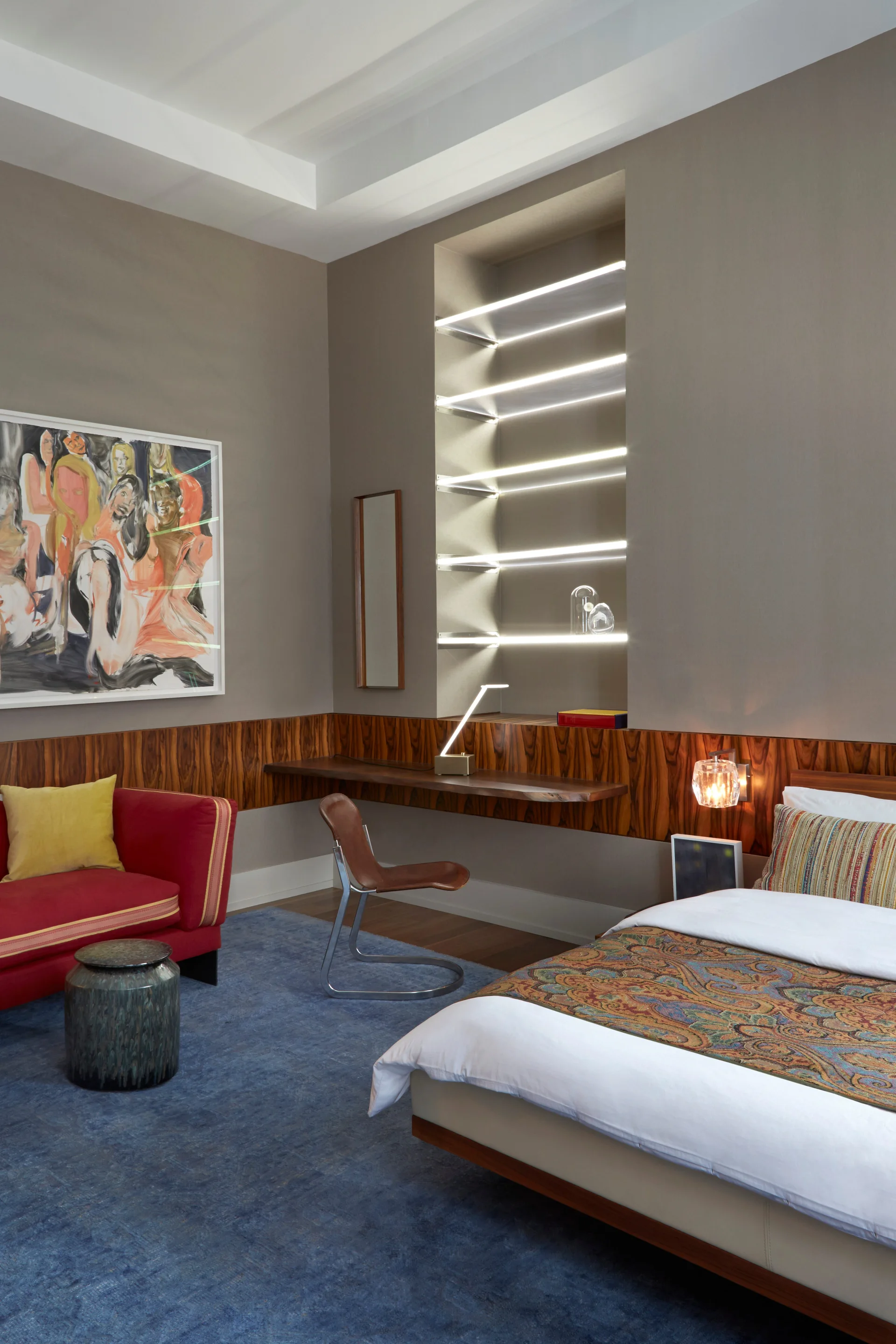 A contemporary bedroom features a floating wooden desk, integrated lighted shelving, a red sofa with a yellow pillow, and a large blue rug.