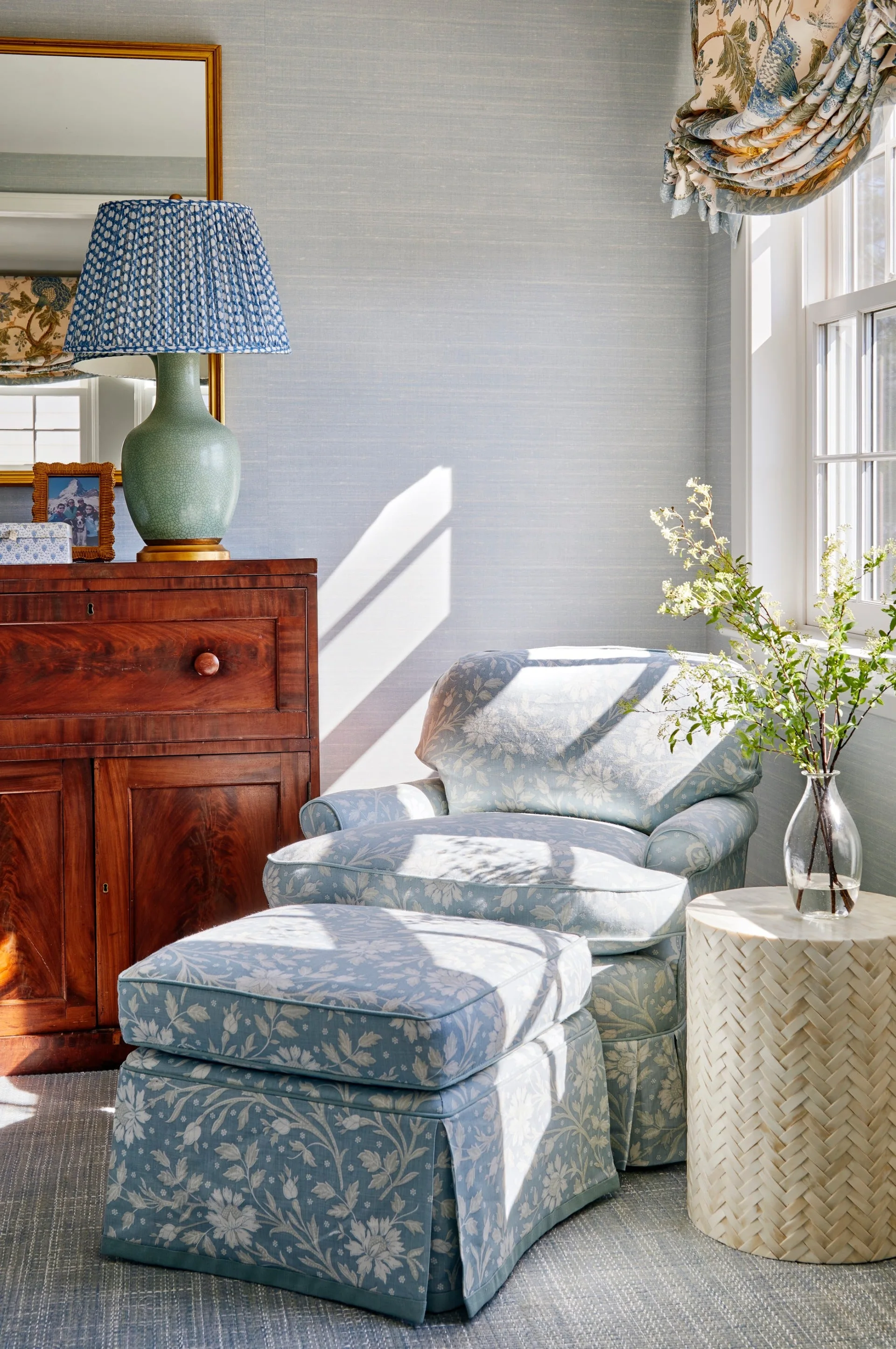 A blue floral armchair and matching ottoman sit next to a dark wood chest of drawers and a woven side table under natural window light.