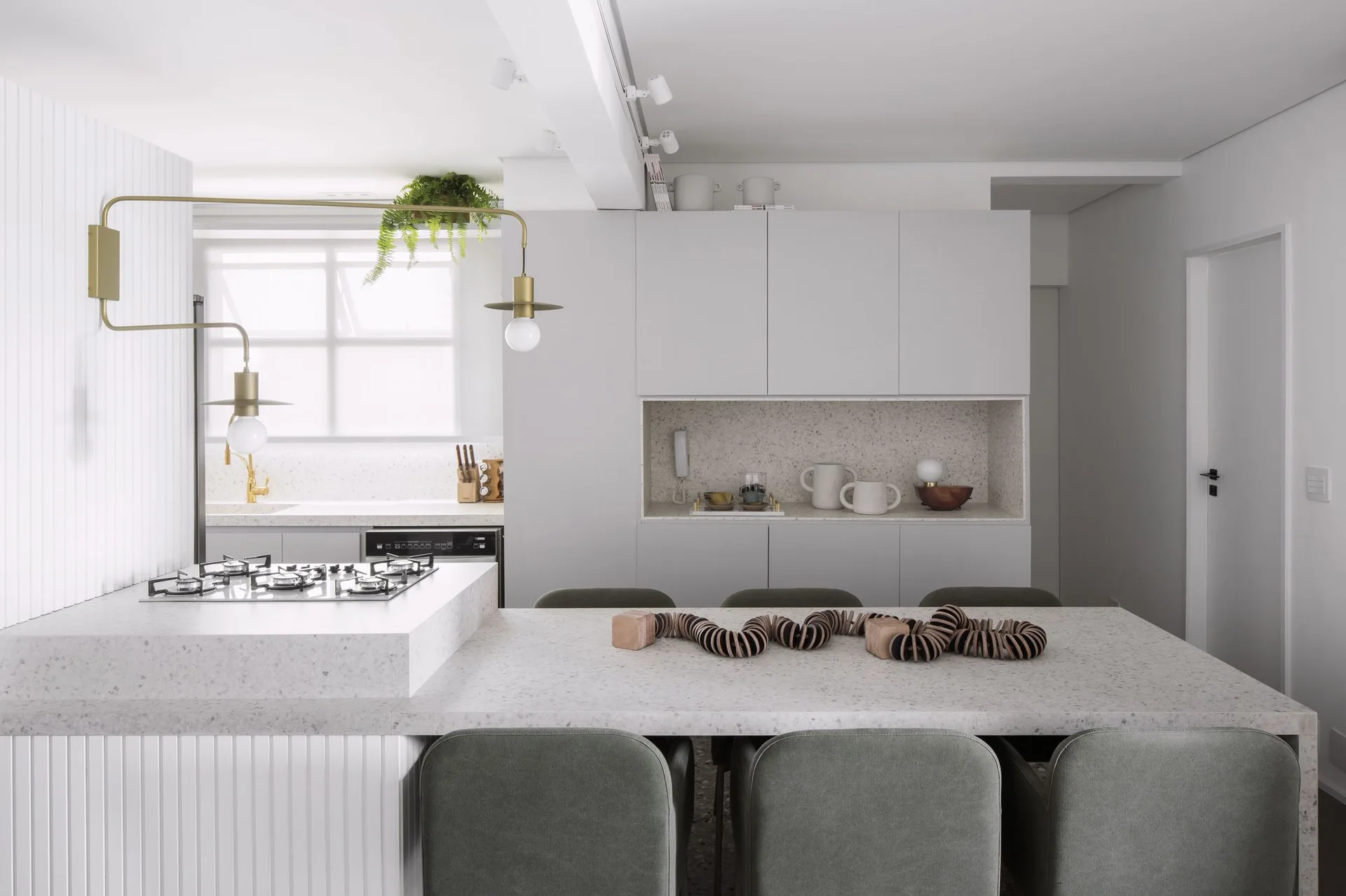 A minimalist kitchen features a light grey island with a speckled stone countertop, matching dining table, green chairs, and a brass swing-arm wall light.