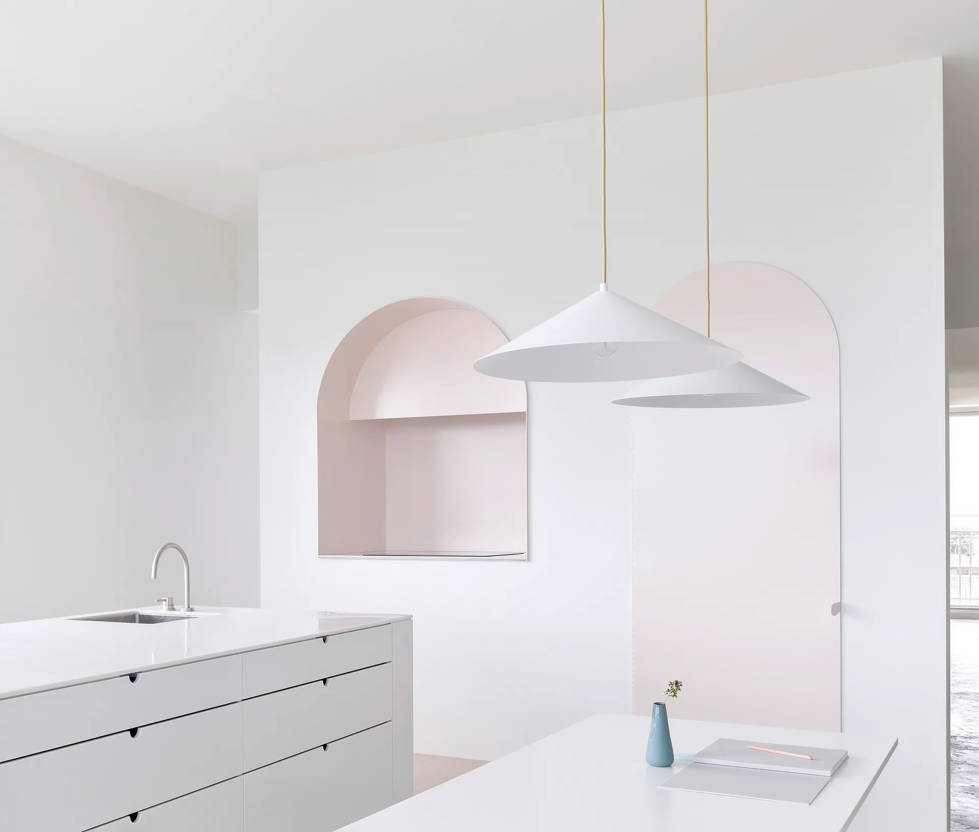A minimalist white kitchen featuring a kitchen island, modern cone pendant lights, and two soft pink arched wall recesses in the background.