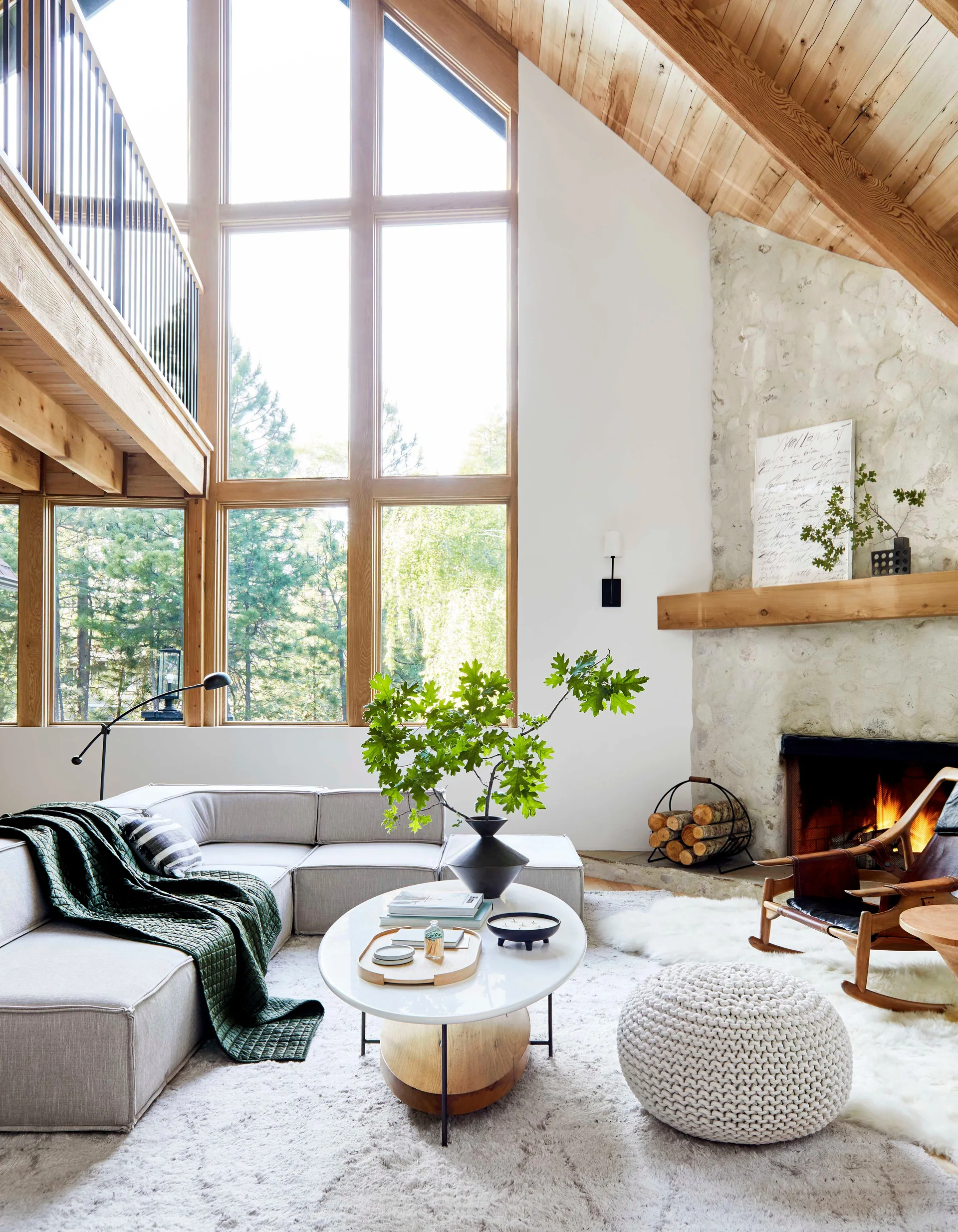 A light-filled living room features a stone fireplace, modular grey sofa, high vaulted wood ceiling, and a large multi-paned window.