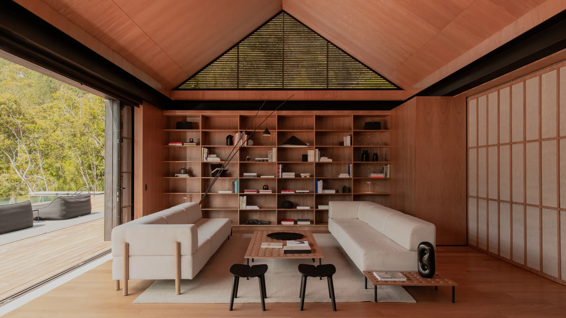 A modern living room features two white sofas, a low wooden coffee table, and a wall of built-in bookshelves beneath a vaulted wooden ceiling.