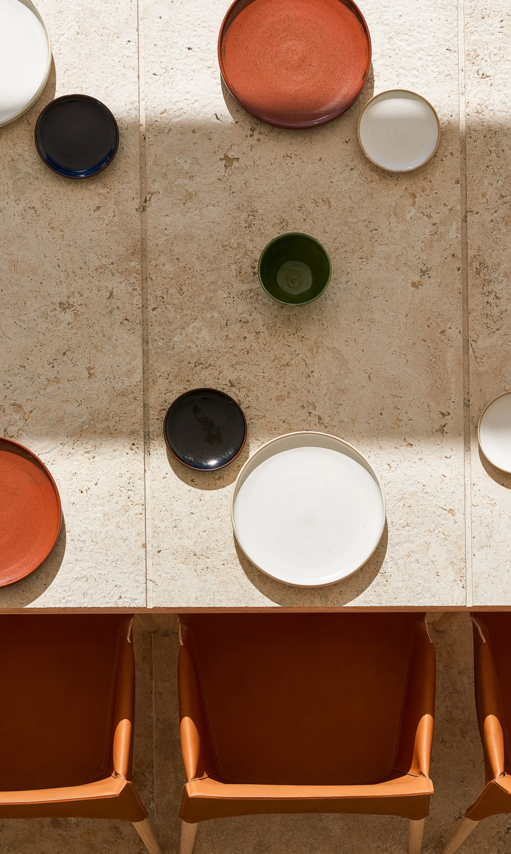An overhead view of a textured beige stone dining table set with colorful ceramic plates and brown leather chairs.