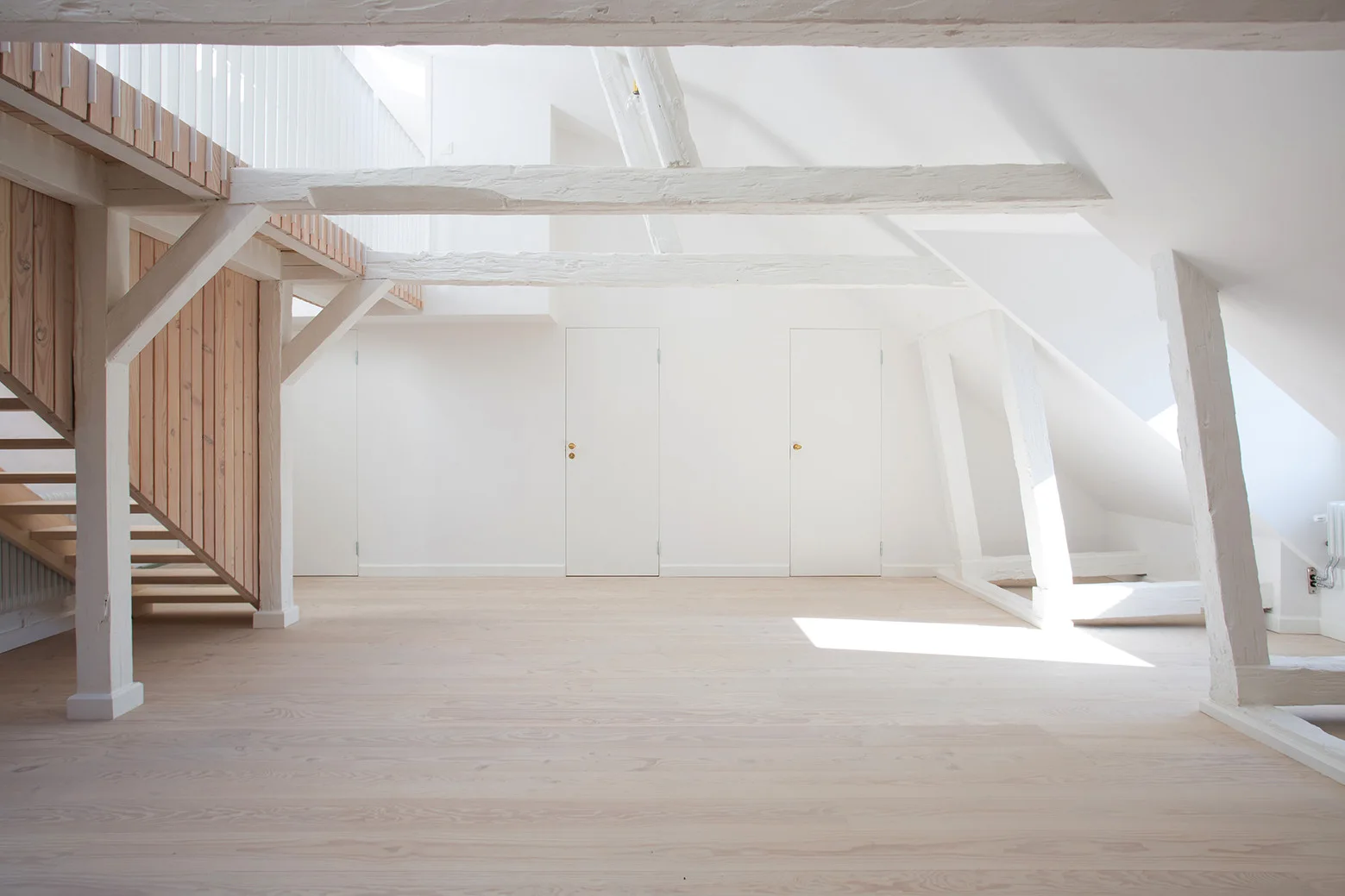 An empty attic landing with white painted walls, exposed ceiling beams, pale wood flooring, and a wooden staircase.