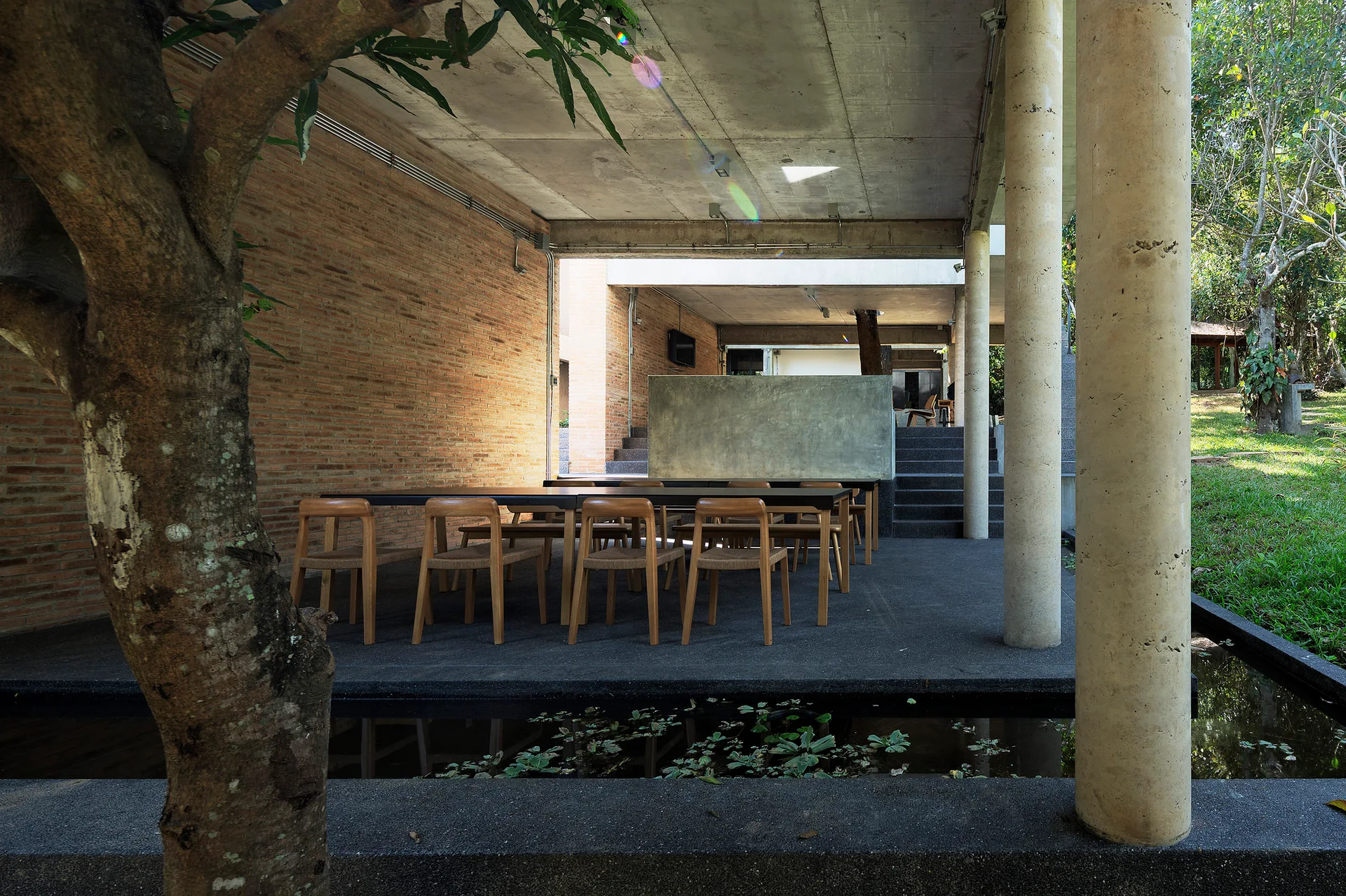 A covered outdoor patio featuring a long wooden dining table and chairs set against an exposed brick wall, concrete columns, and a small foreground pond.