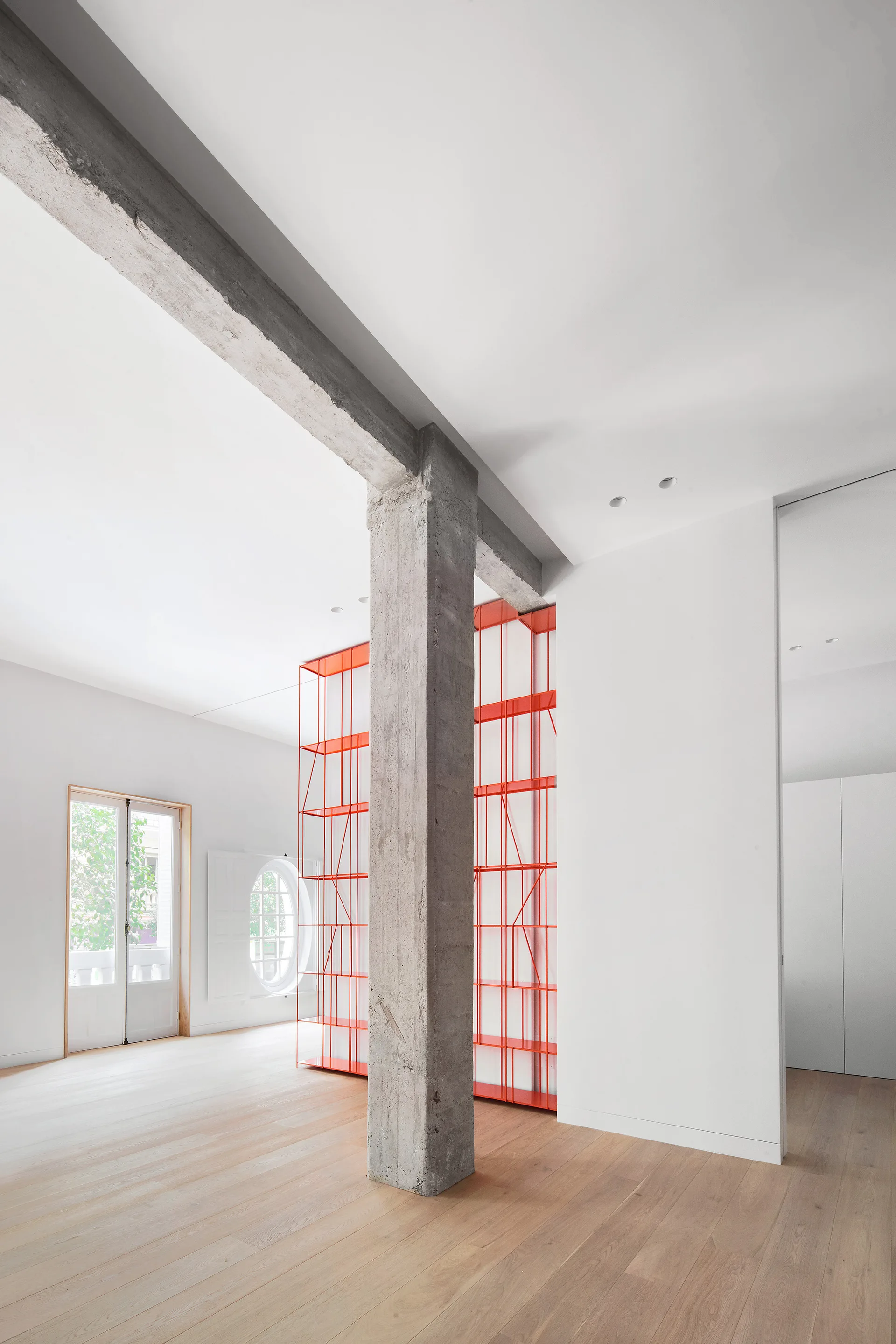 A minimalist room featuring light wood flooring, white walls, an exposed concrete column, and a tall red metal wire shelving unit.
