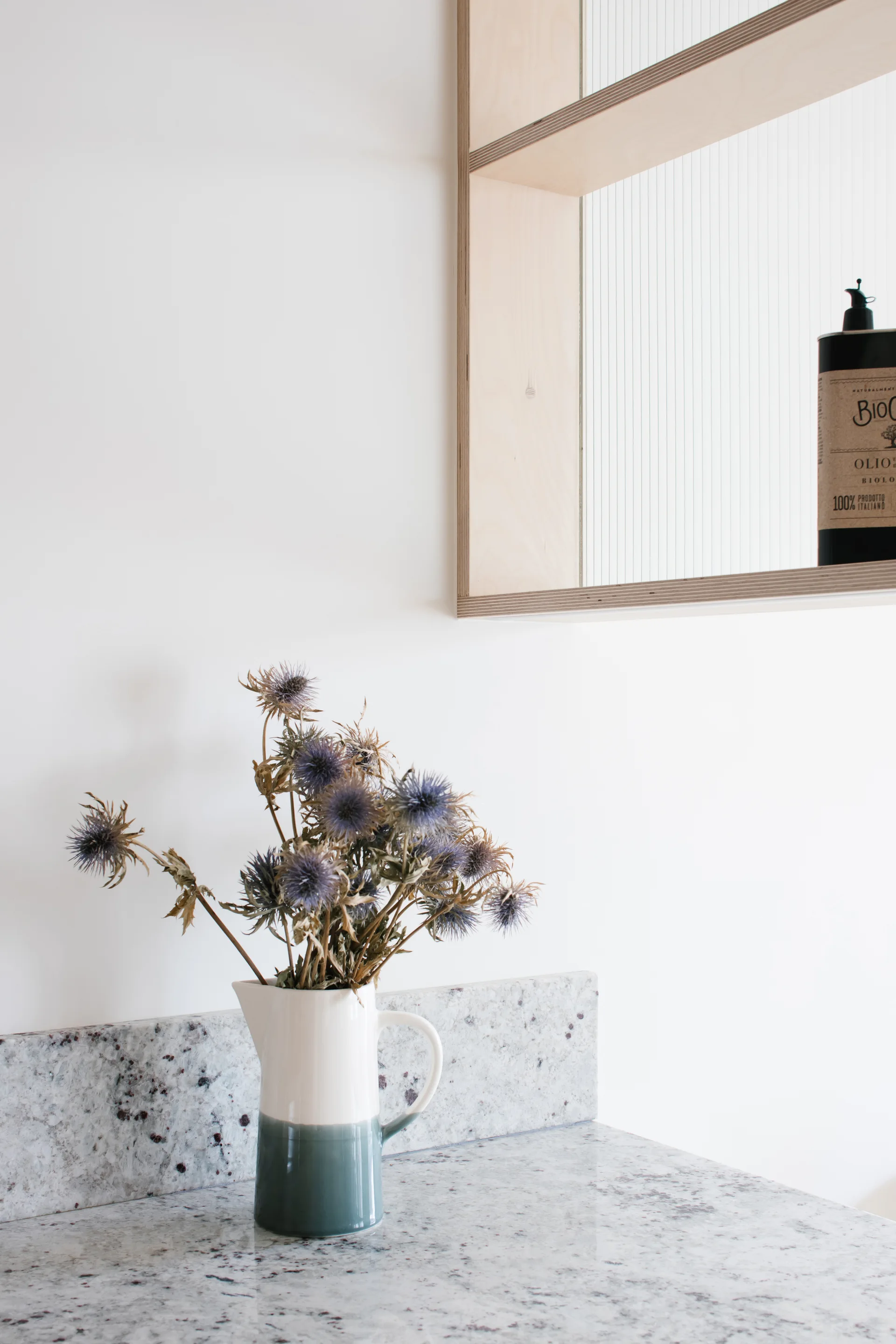 A speckled granite countertop and matching short backsplash hold a two-tone ceramic pitcher filled with dried thistle flowers beneath a light plywood shelf.