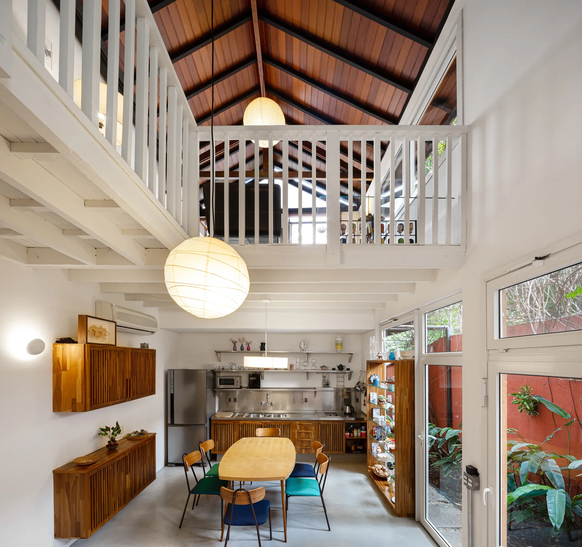 A dining space with an oval wooden table and colorful chairs beneath a vaulted wood-plank ceiling and a white mezzanine railing.
