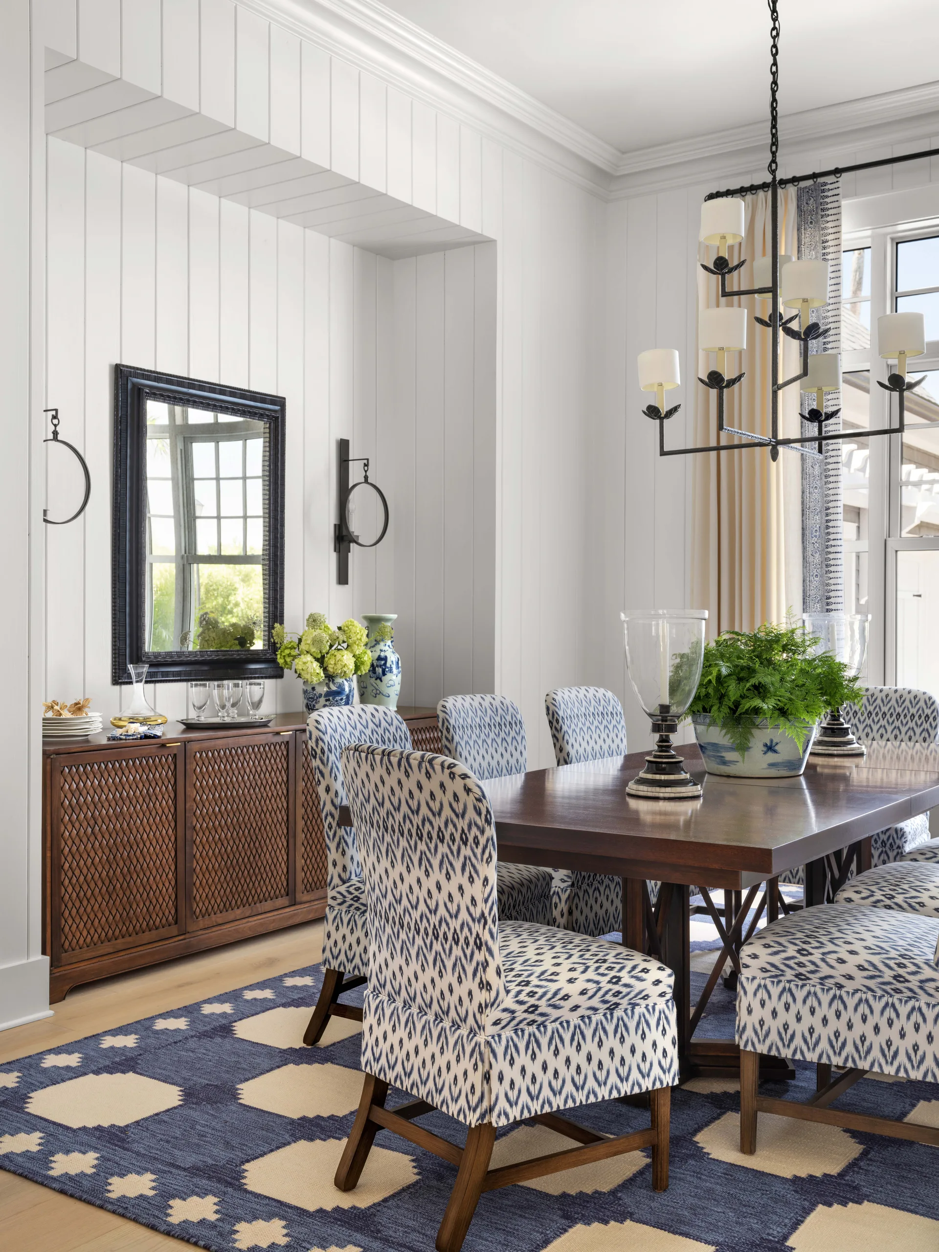 A dining room features blue and white ikat upholstered chairs around a dark wood table on a geometric rug, set against vertical white paneled walls.