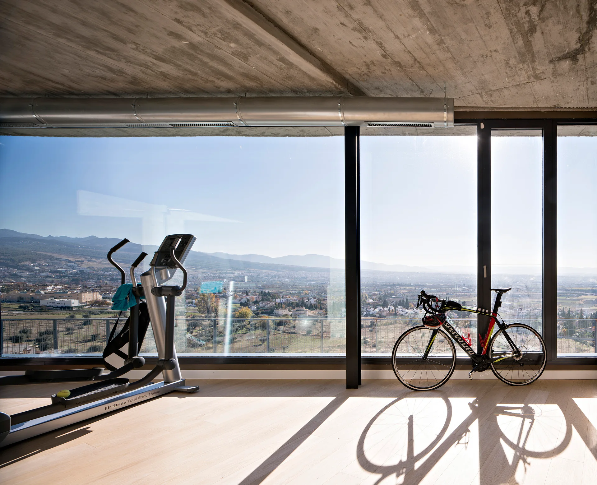 A minimalist home gym featuring light wood flooring, an elliptical machine, a road bike, and an exposed concrete ceiling facing large windows.