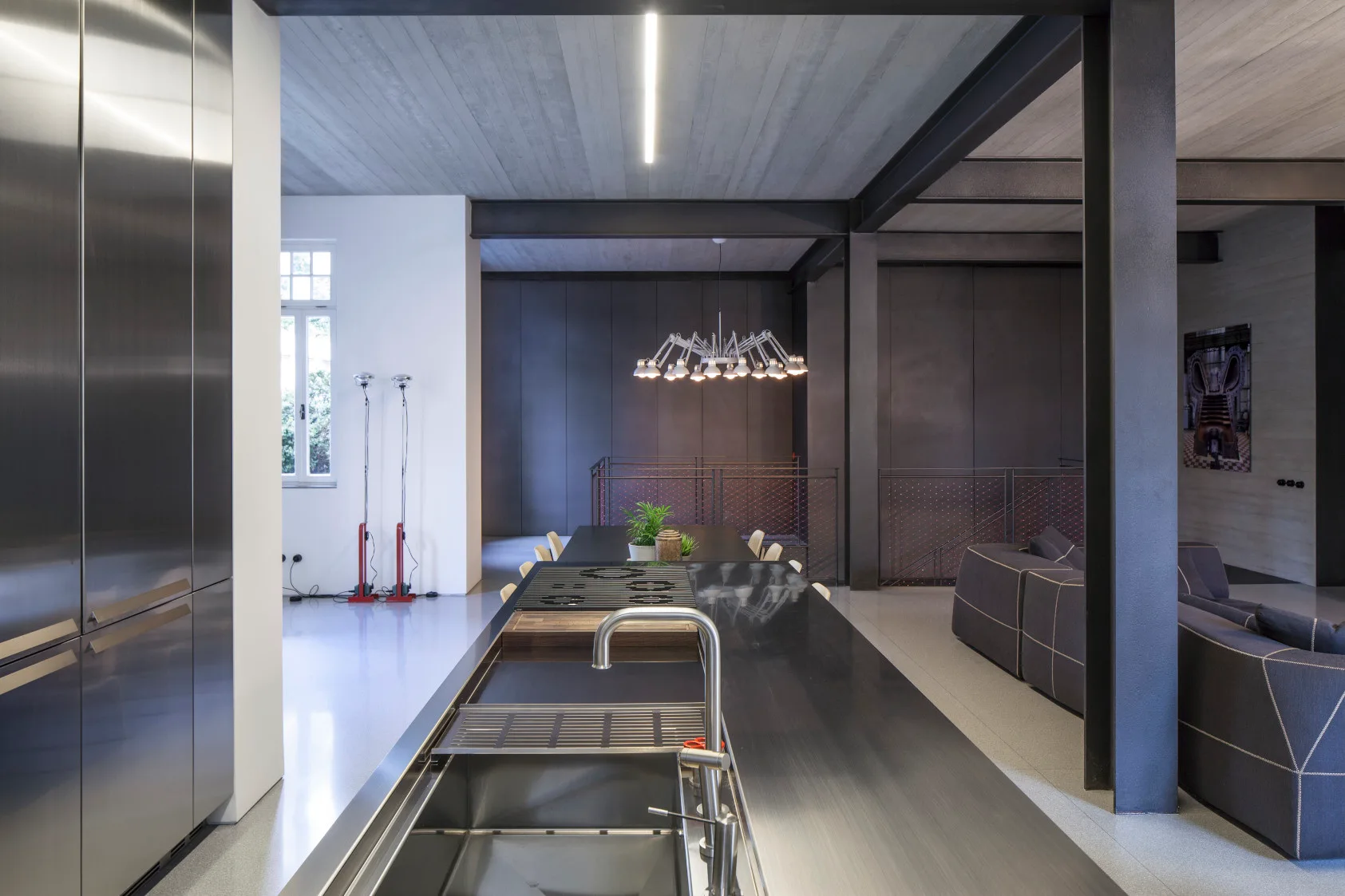 A spacious industrial loft kitchen features a long stainless steel island, exposed black steel beams, and a gray concrete ceiling.