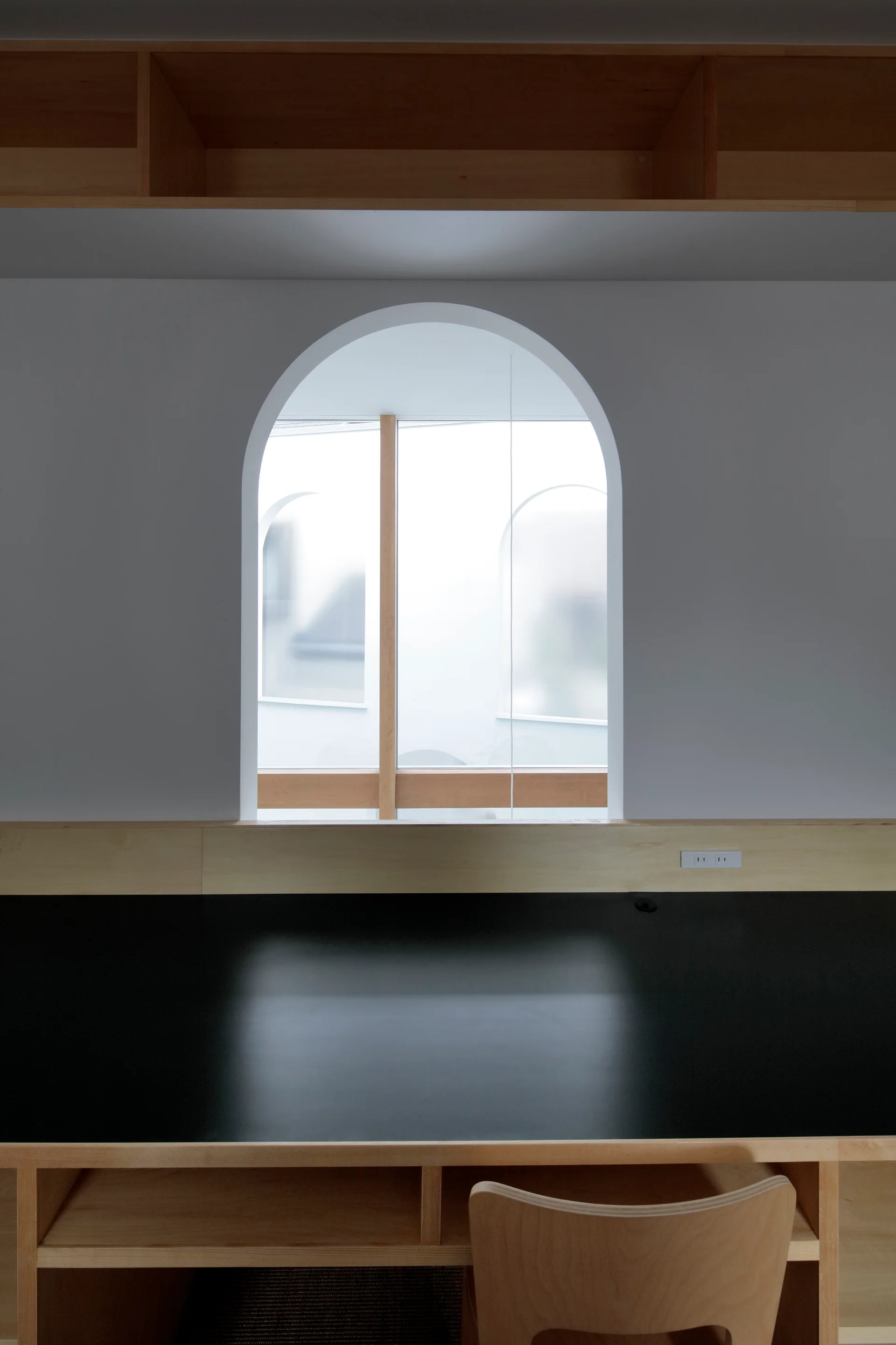 A minimalist black desk with a light wood chair sits below a white wall featuring a prominent arched window and overhead wooden shelving.