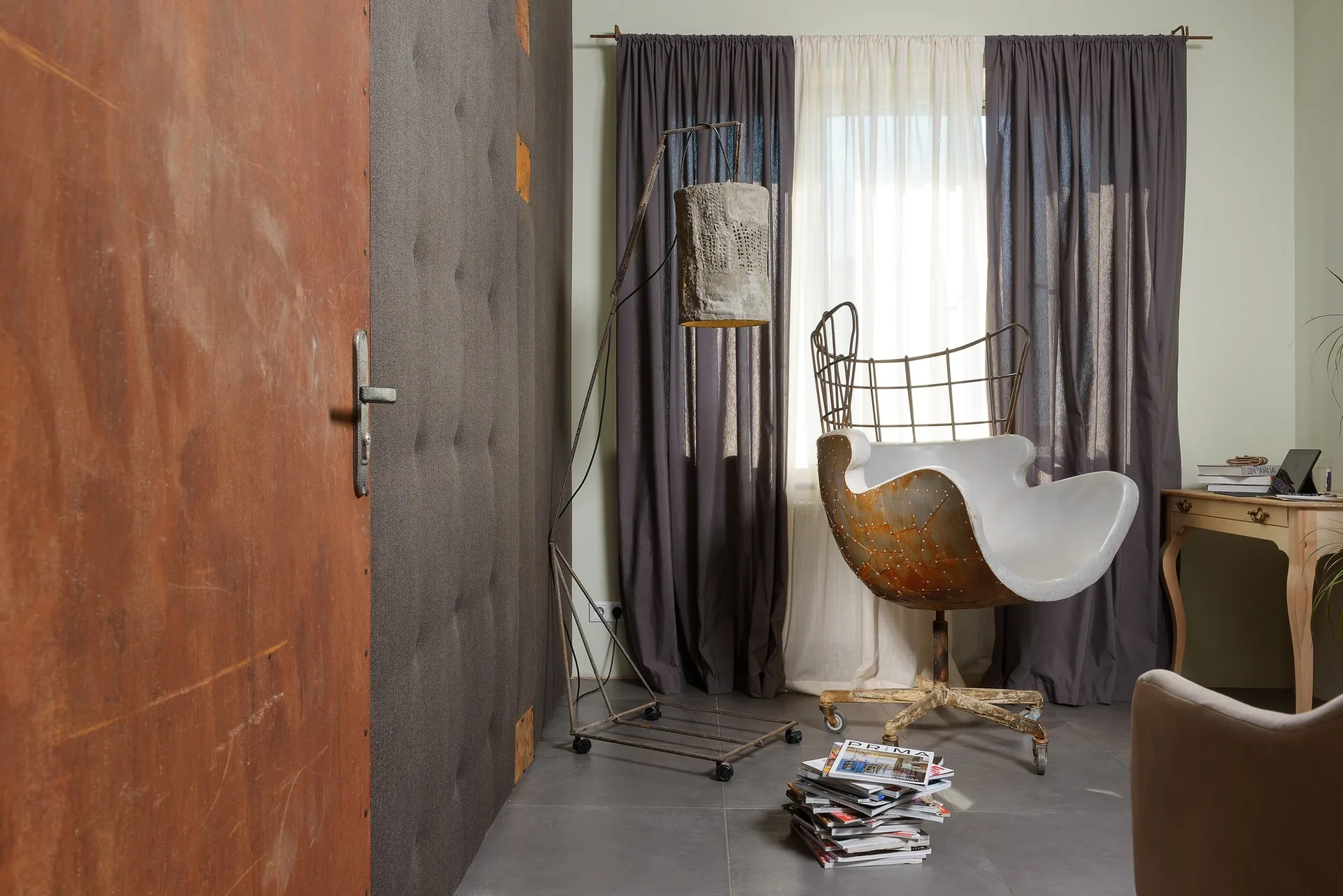 An industrial seating area features a rusted metal armchair, a rebar floor lamp, and a dark grey tufted wall panel.