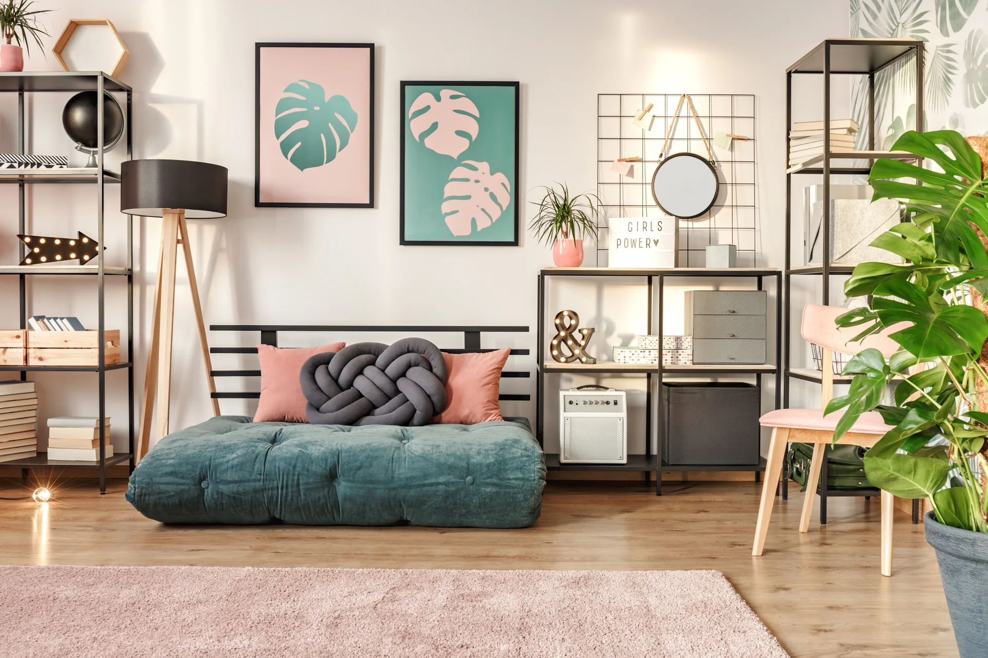 A low green tufted sofa sits on a wood floor next to black metal shelves, a pink rug, and framed tropical leaf prints.