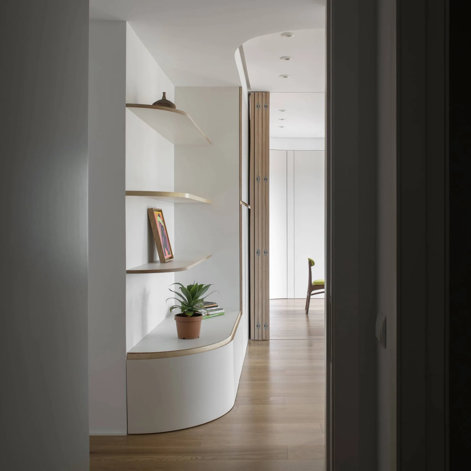 A modern hallway features a curved white built-in shelving unit with light wood edges, leading toward a brightly lit room.