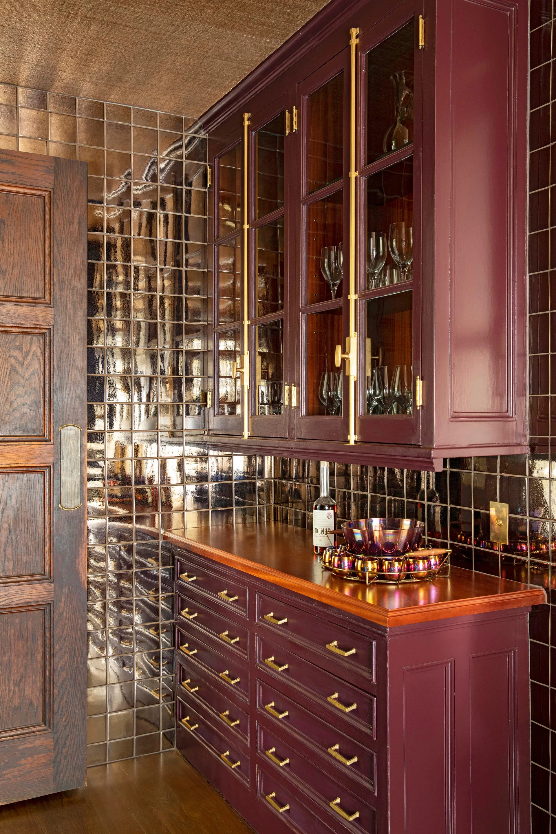 Burgundy cabinets with brass hardware and a wooden countertop are set against a reflective, metallic square tile backsplash.