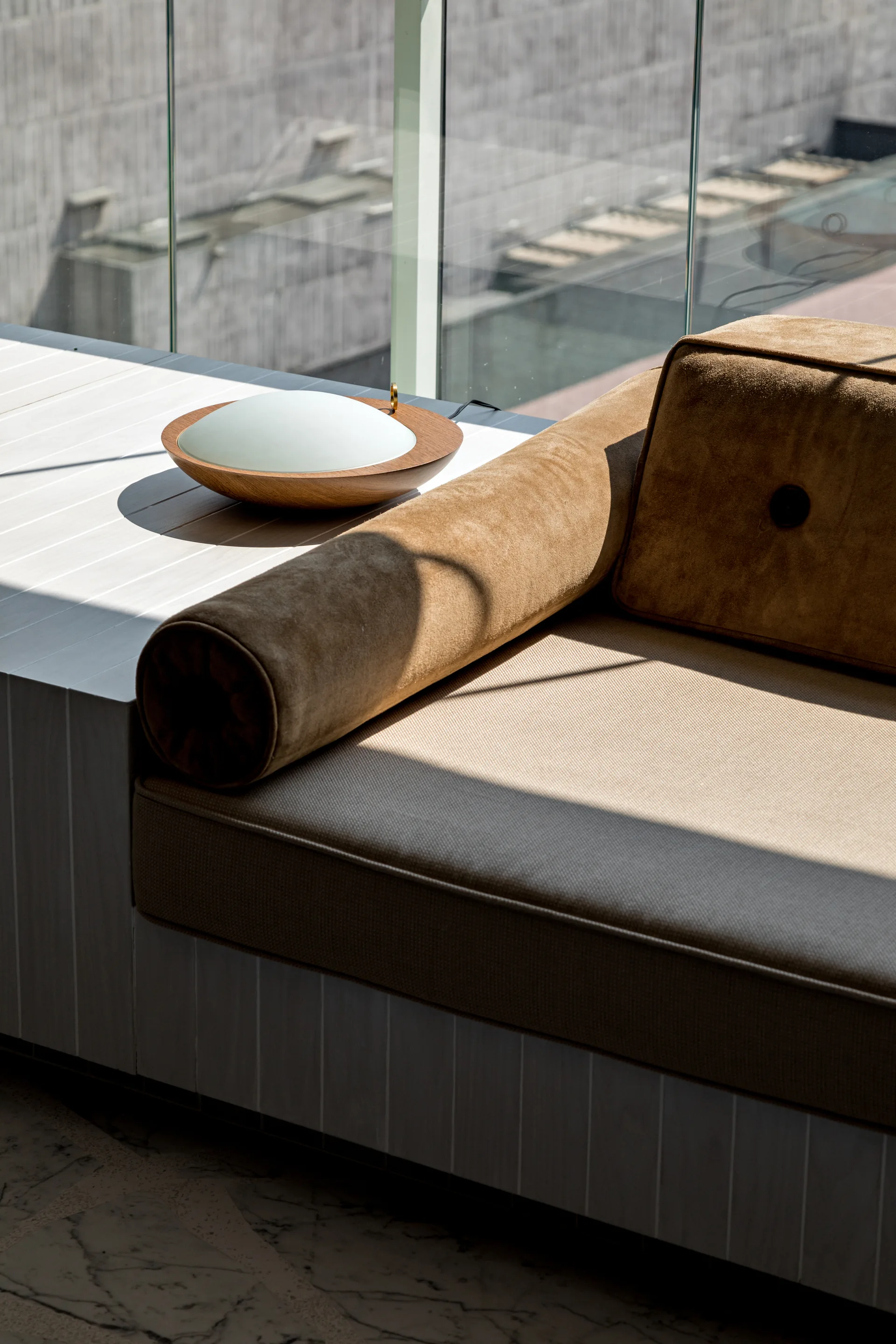 A close-up of a tan upholstered daybed with a bolster cushion and a circular wood lamp resting on a white vertical-slat built-in bench.