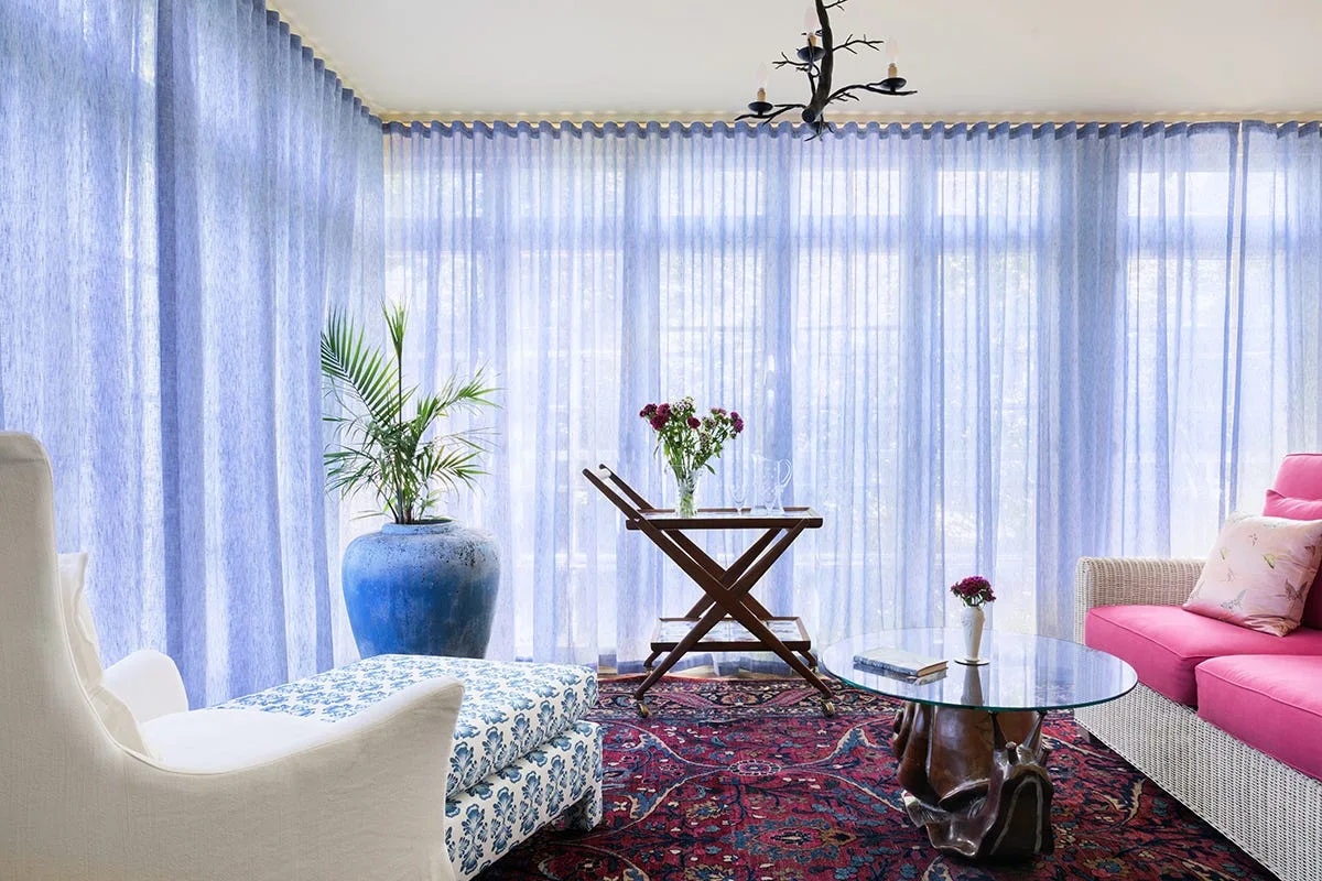 A sunlit eclectic living room with sheer blue curtains draped across large windows, a pink sofa, a patterned ottoman, and a red oriental rug.