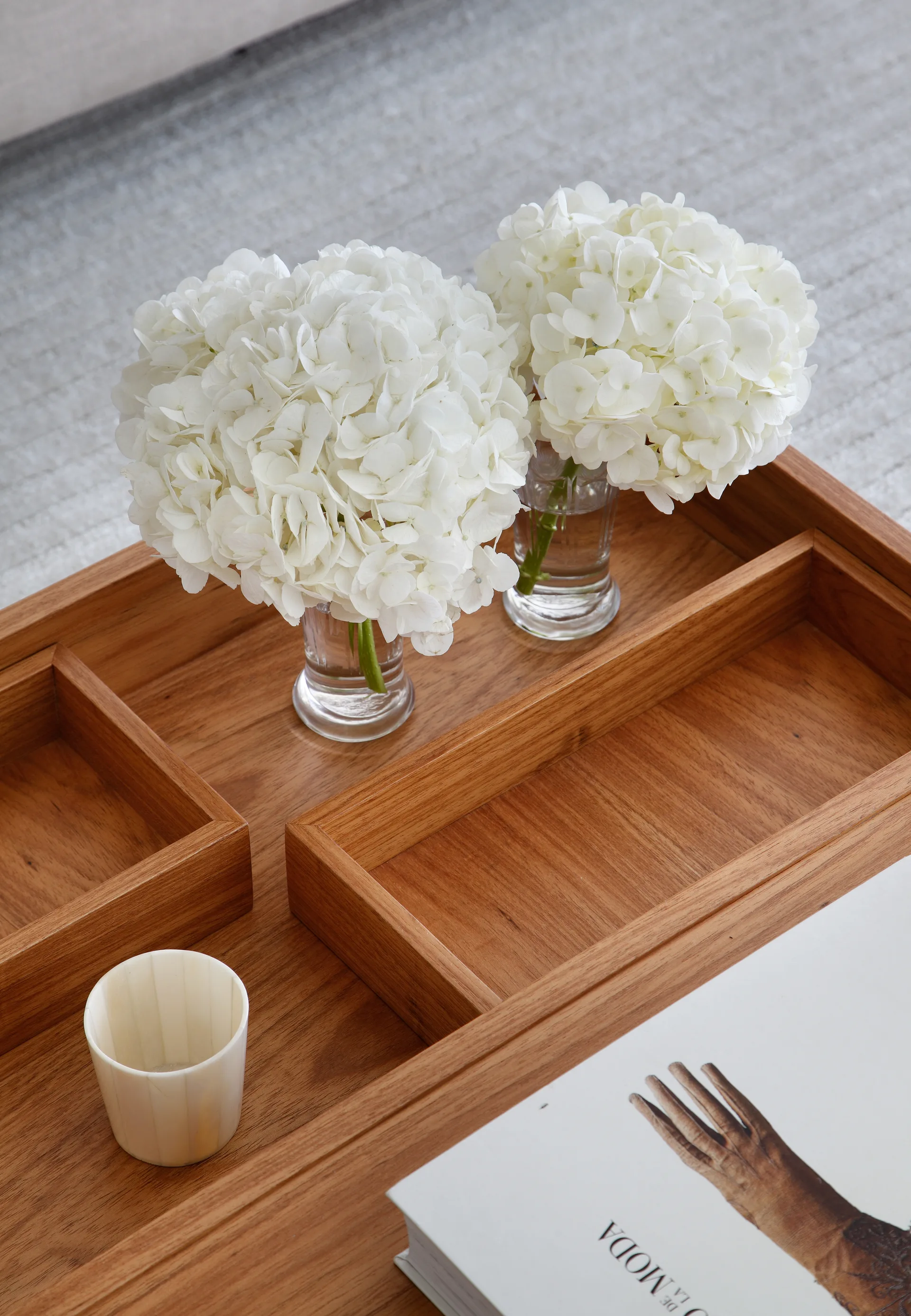 A compartmentalized wooden tray holds two clear glass vases with white hydrangea blooms, a small textured cream cup, and a book.