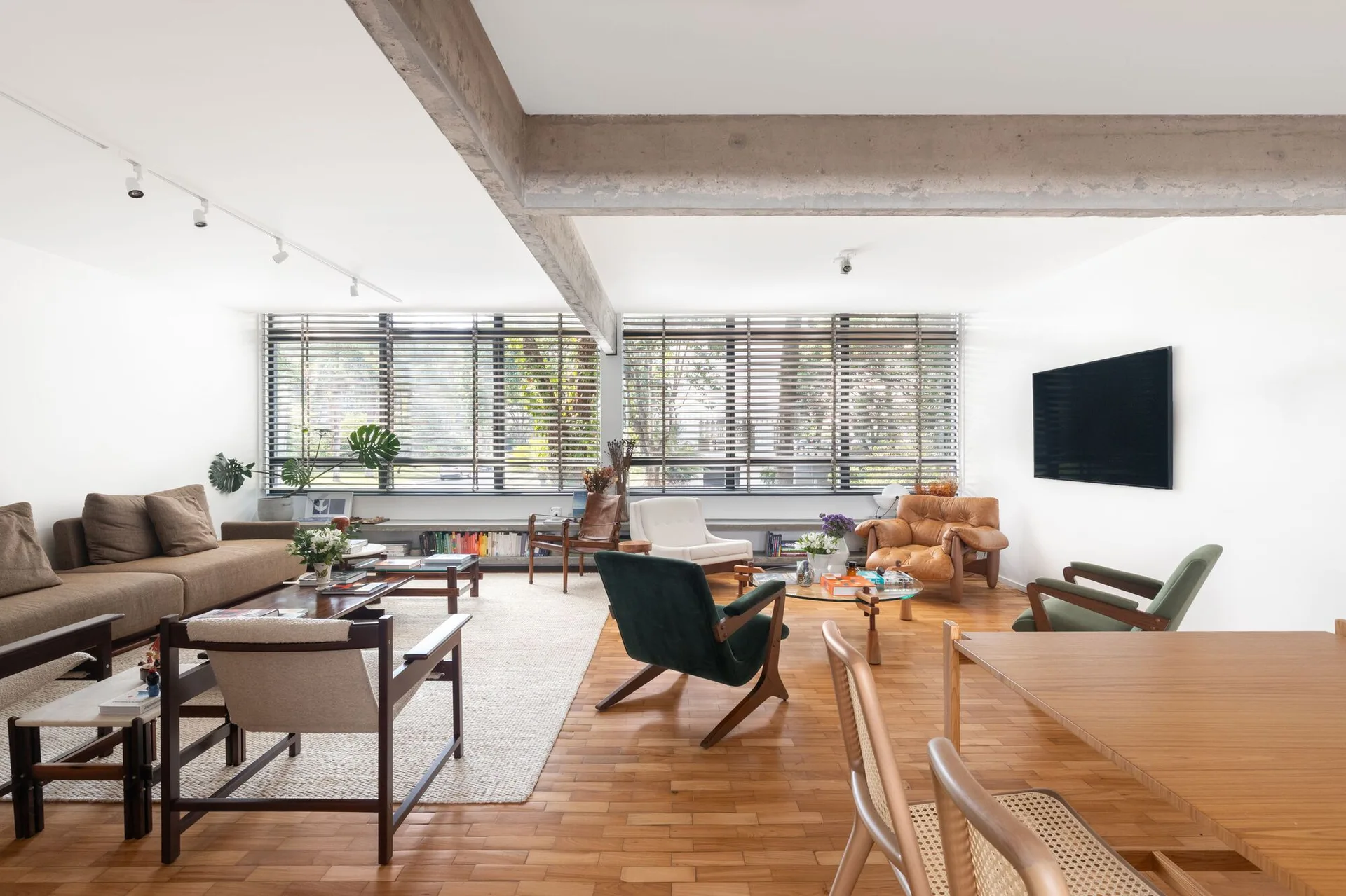 Mid-century modern living room featuring warm wood parquet flooring, a concrete ceiling beam, and various wood and leather armchairs near large windows.