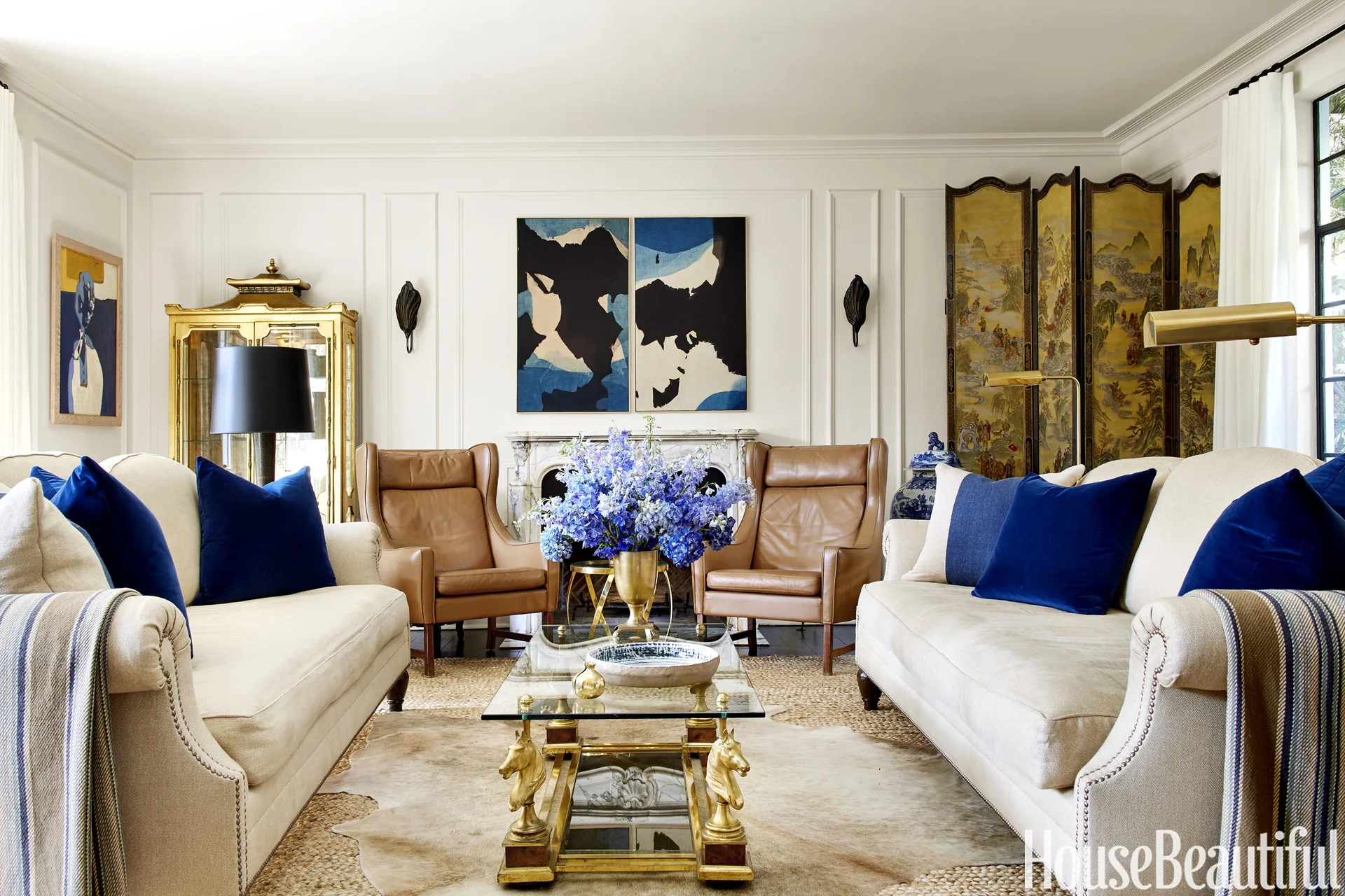 A symmetrical living room with two off-white sofas, cobalt blue pillows, tan leather armchairs, and a glass coffee table in front of a fireplace.
