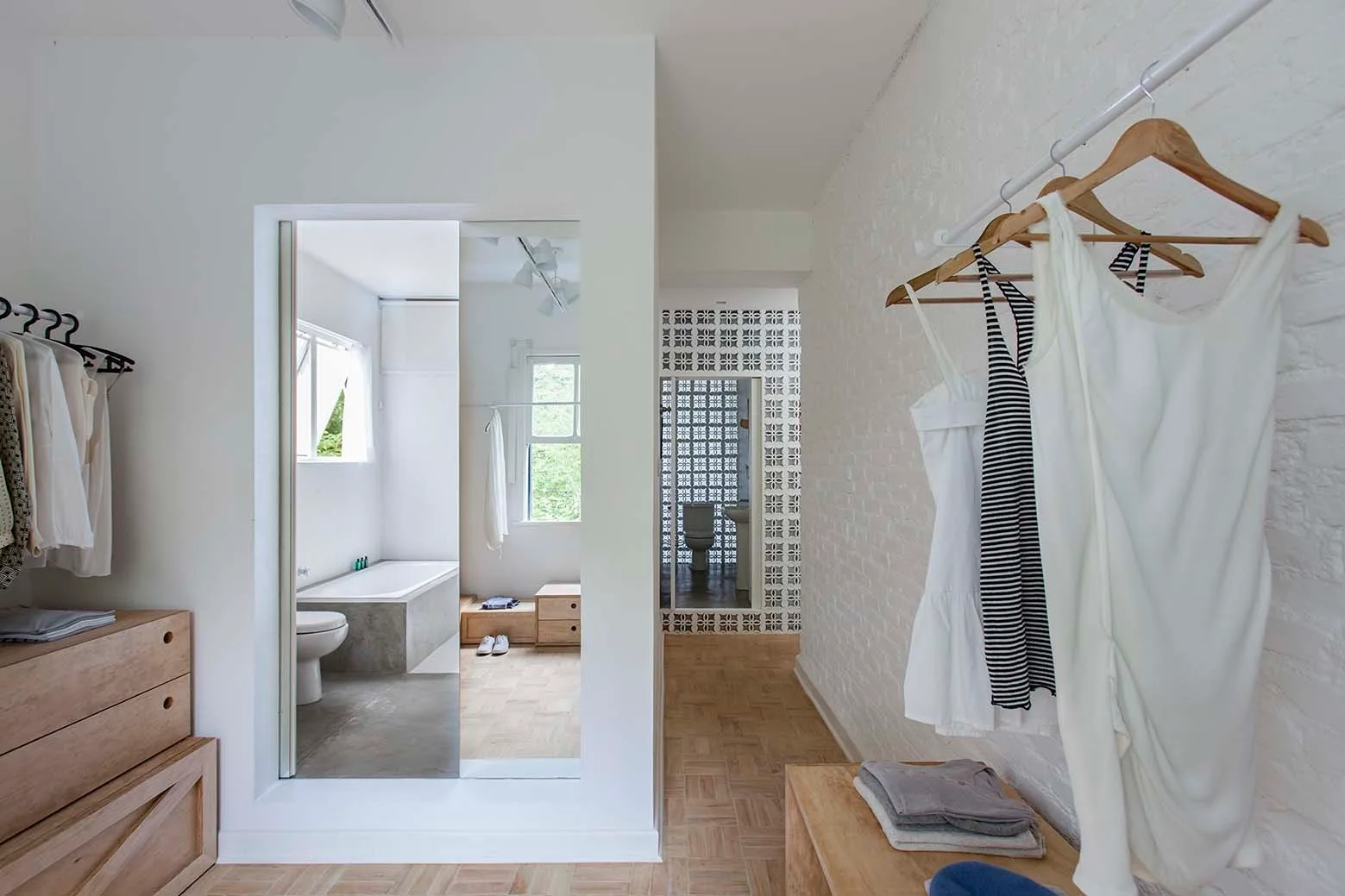 A minimalist dressing room features white painted brick walls, wooden clothing hangers on a rack, light oak drawers, and a view into a bright bathroom.
