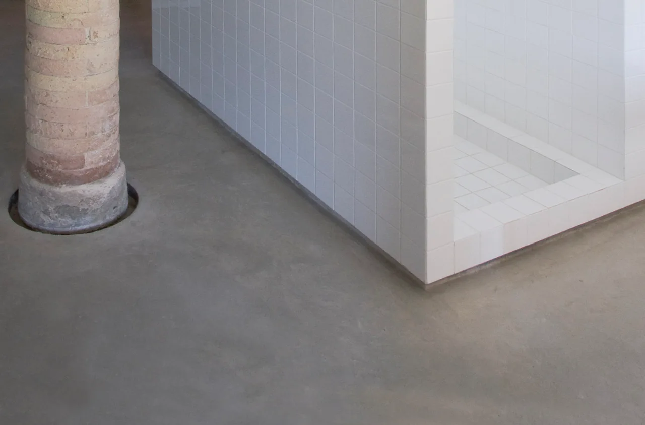 A white grid-tiled shower enclosure stands on a smooth grey concrete floor next to an exposed brick column.