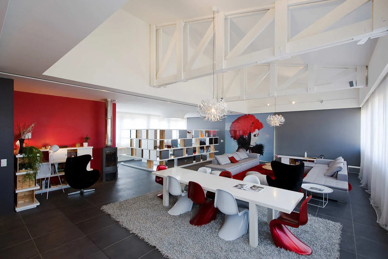 A spacious open-plan living and dining room featuring white exposed ceiling trusses, a red accent wall, and sleek modern furniture.