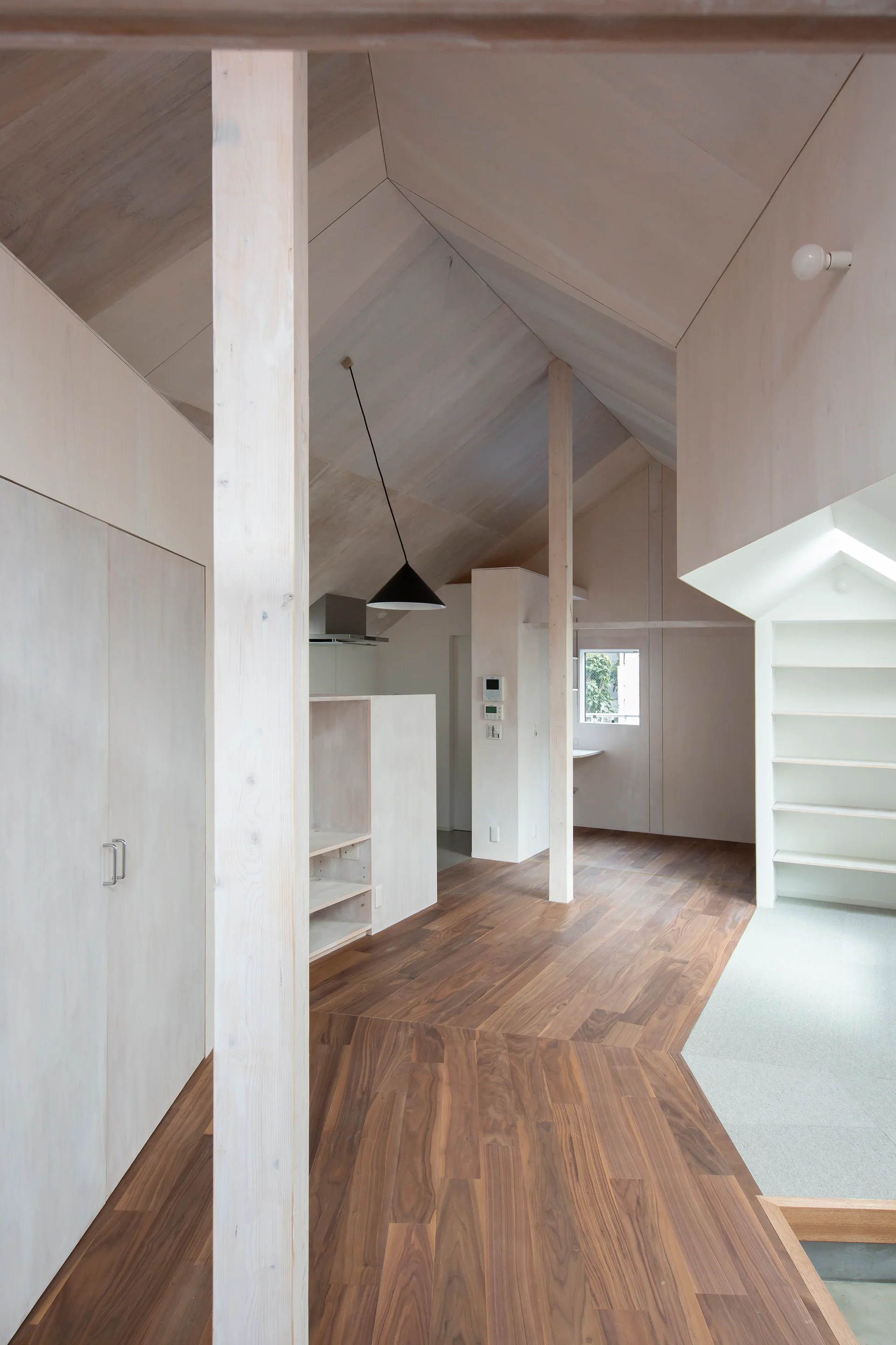 A minimalist residential interior features light wood paneled vaulted ceilings, dark wood plank floors, structural columns, and built-in storage units.