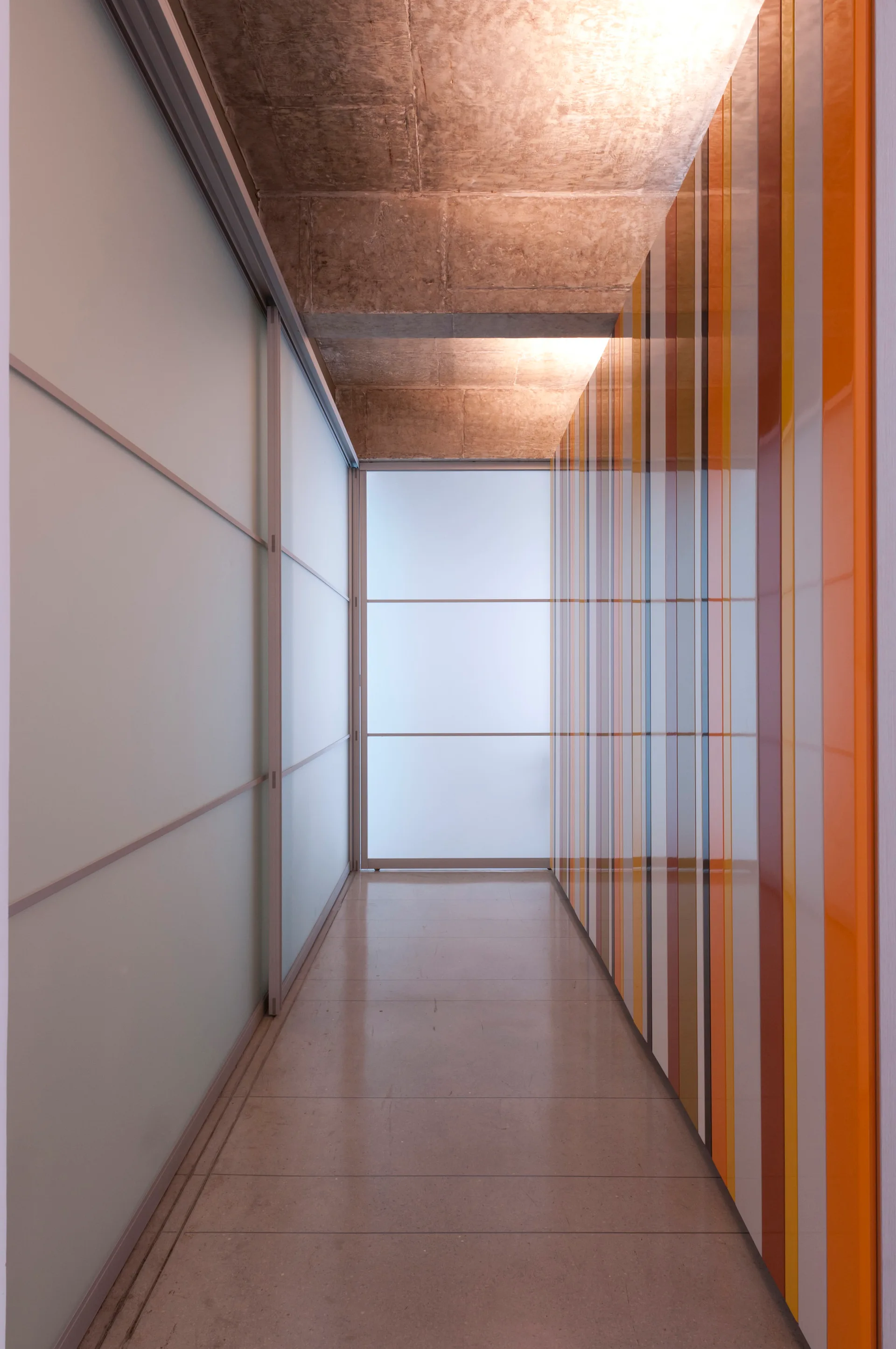 A narrow hallway featuring a glossy multi-colored striped wall, sliding frosted glass doors, and an exposed concrete ceiling.