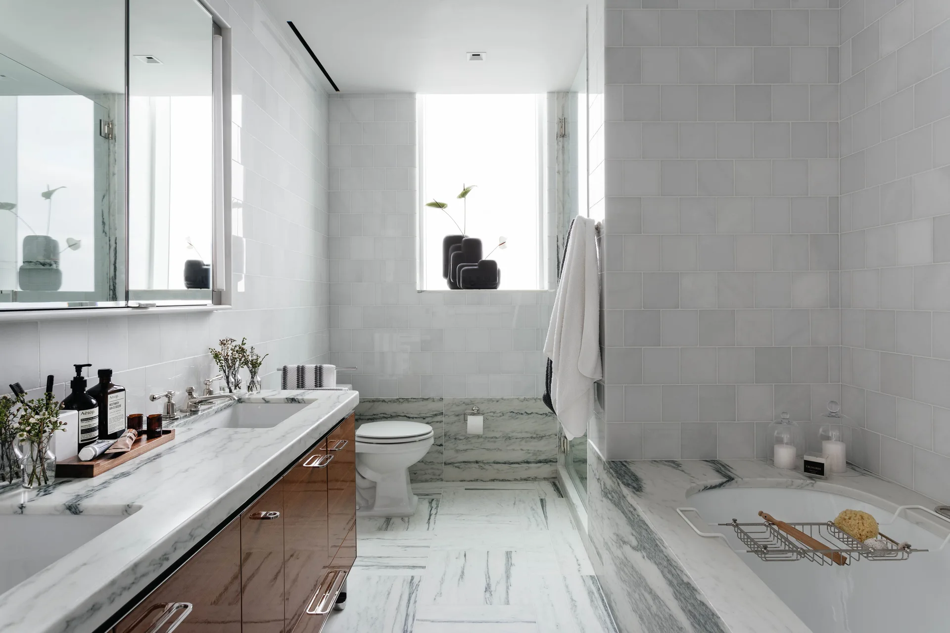 A bright bathroom features a glossy wood vanity with a marble countertop, white subway tile walls, and a large marble-clad bathtub.