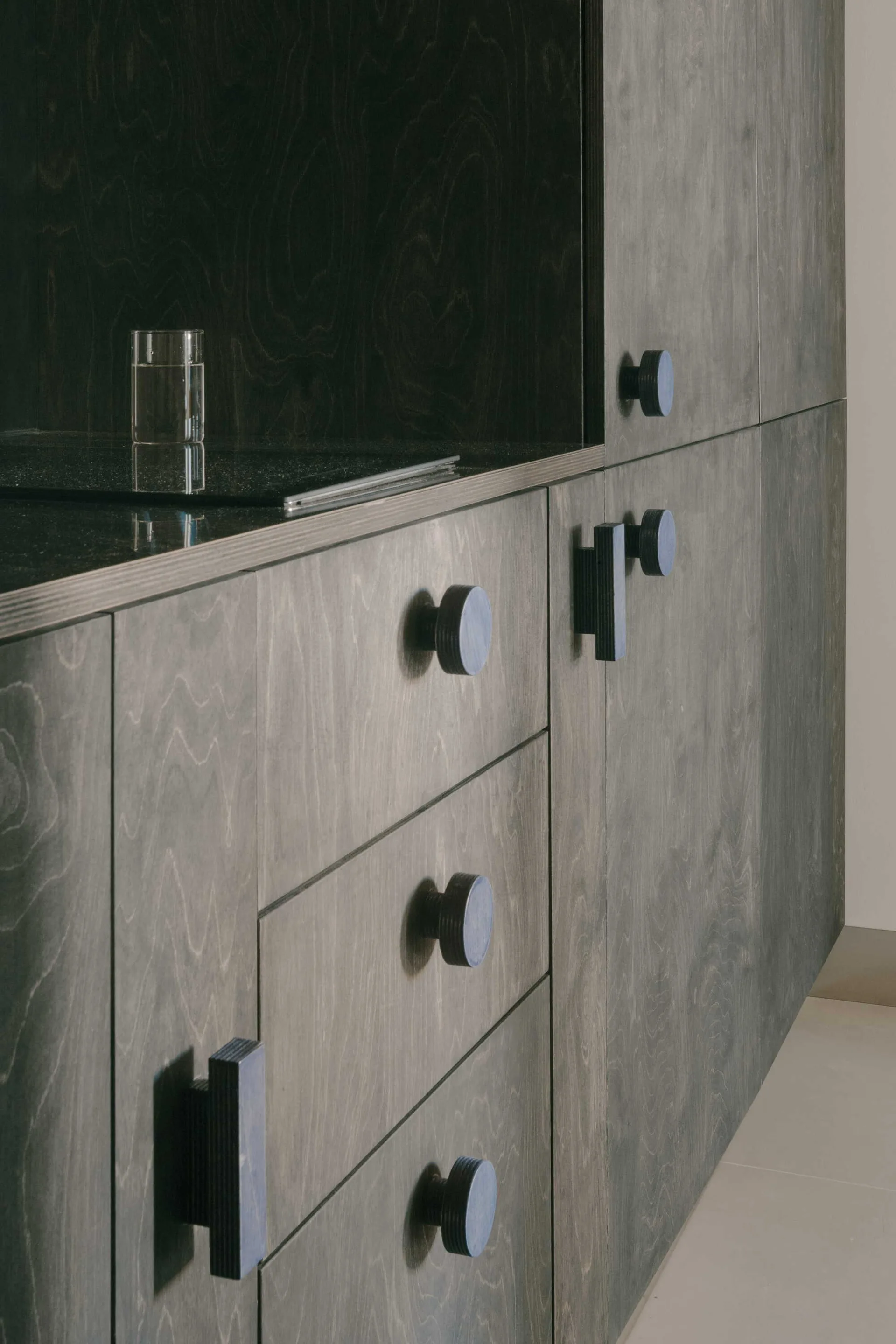 Close-up of charcoal grey stained plywood kitchen cabinetry featuring thick circular and rectangular wooden knobs and a black countertop.