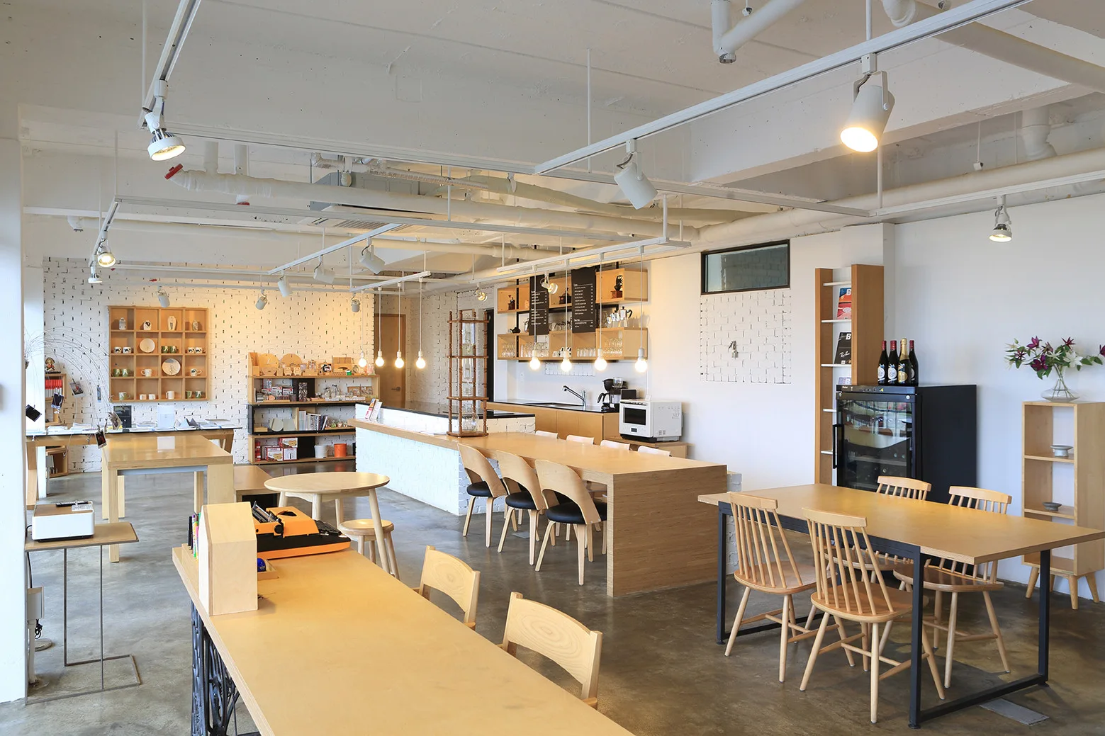 A spacious interior featuring light wood tables and chairs, painted white brick walls, exposed ceiling pipes, and hanging pendant lights.