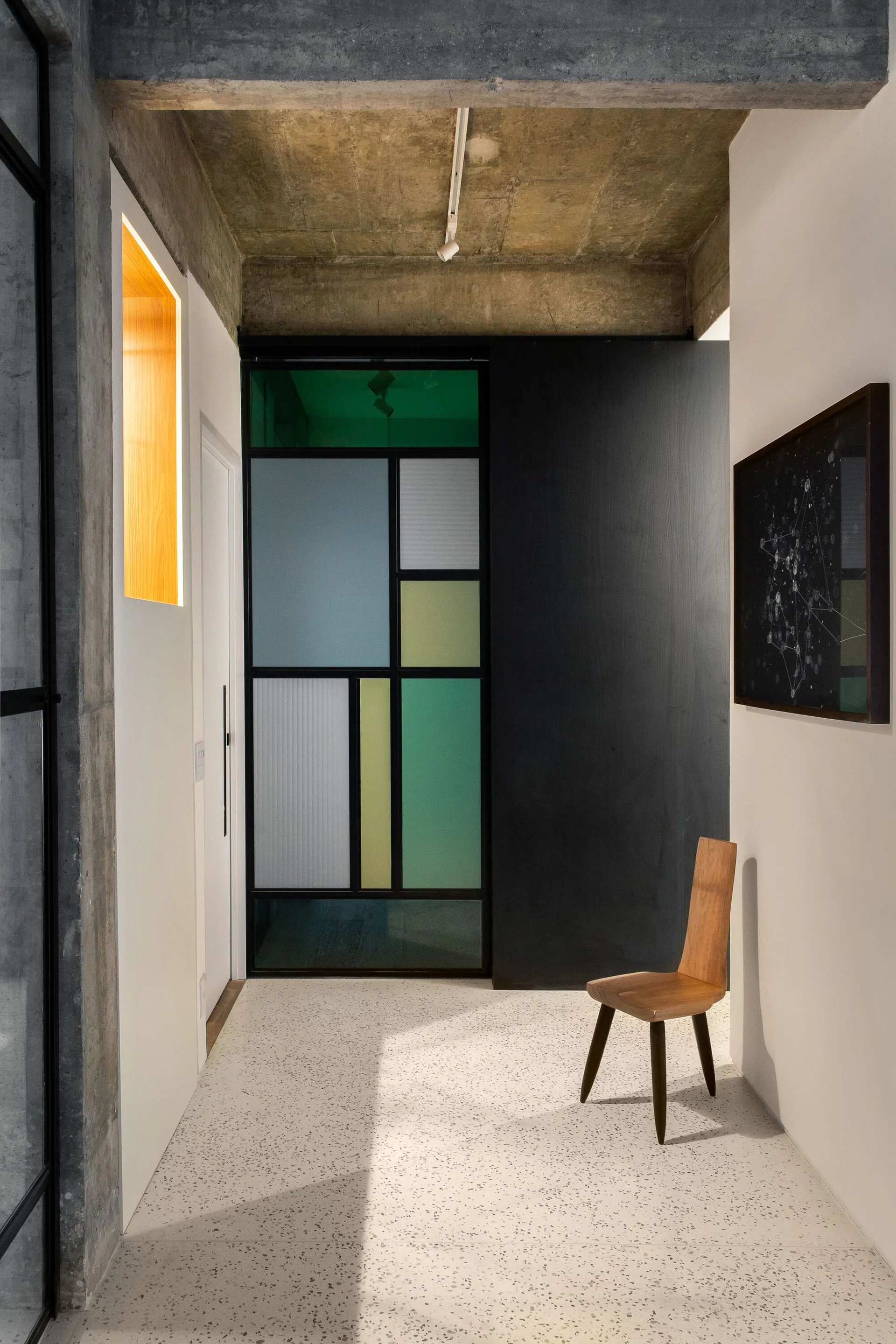 A modern hallway features light speckled terrazzo flooring, a minimalist wood chair, and a geometric partition with multicolored glass panes.