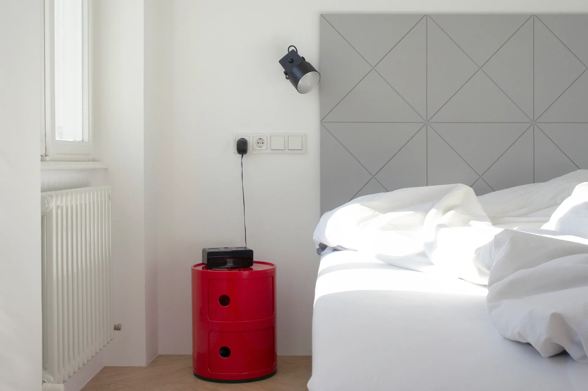 A modern bedroom features a gray geometric patterned headboard, white bedding, a black wall sconce, and a bright red cylindrical nightstand.