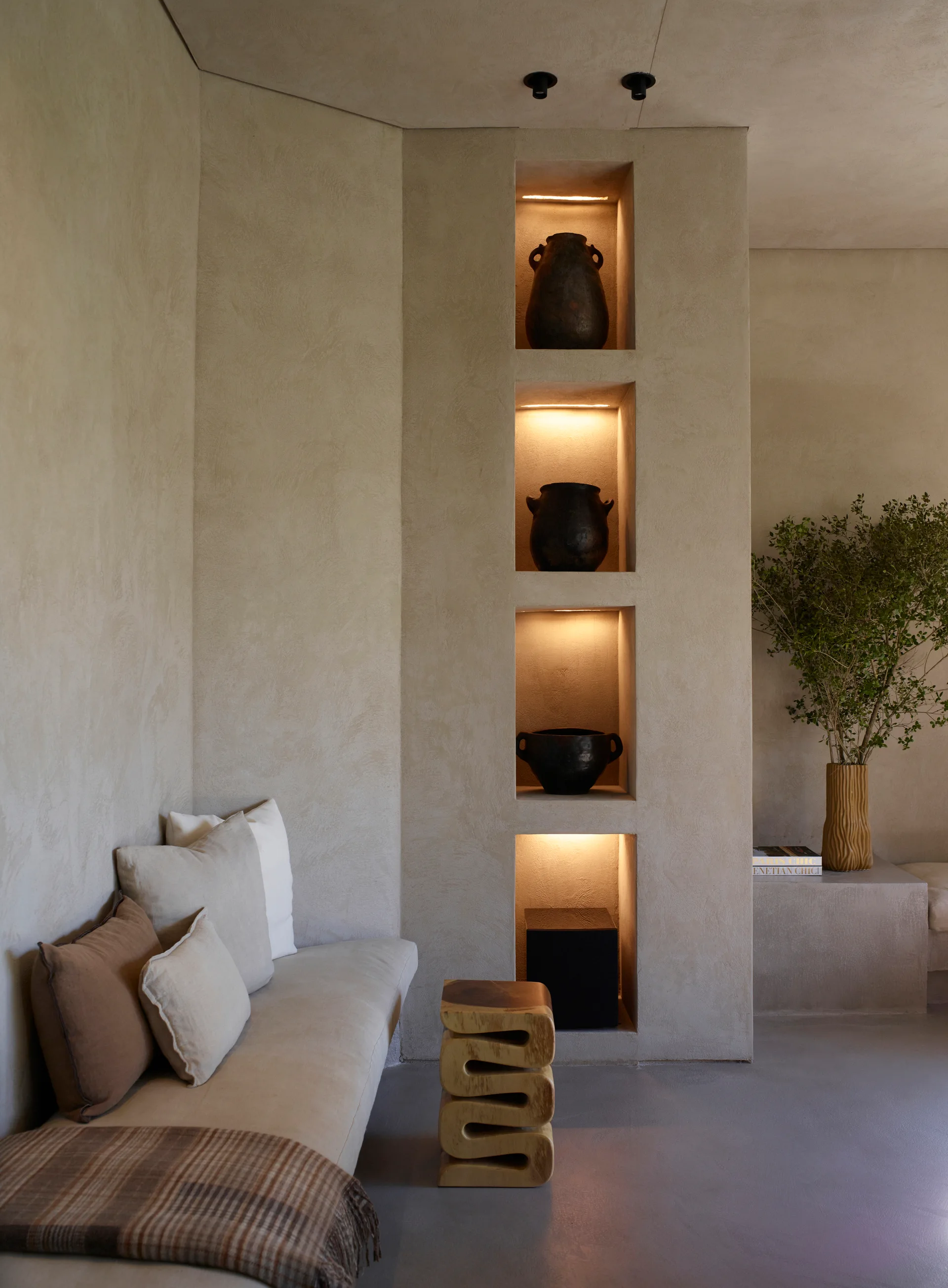 A minimalist plaster lounge area features a built-in bench sofa, a wavy wood side table, and vertical illuminated wall niches displaying dark pottery.