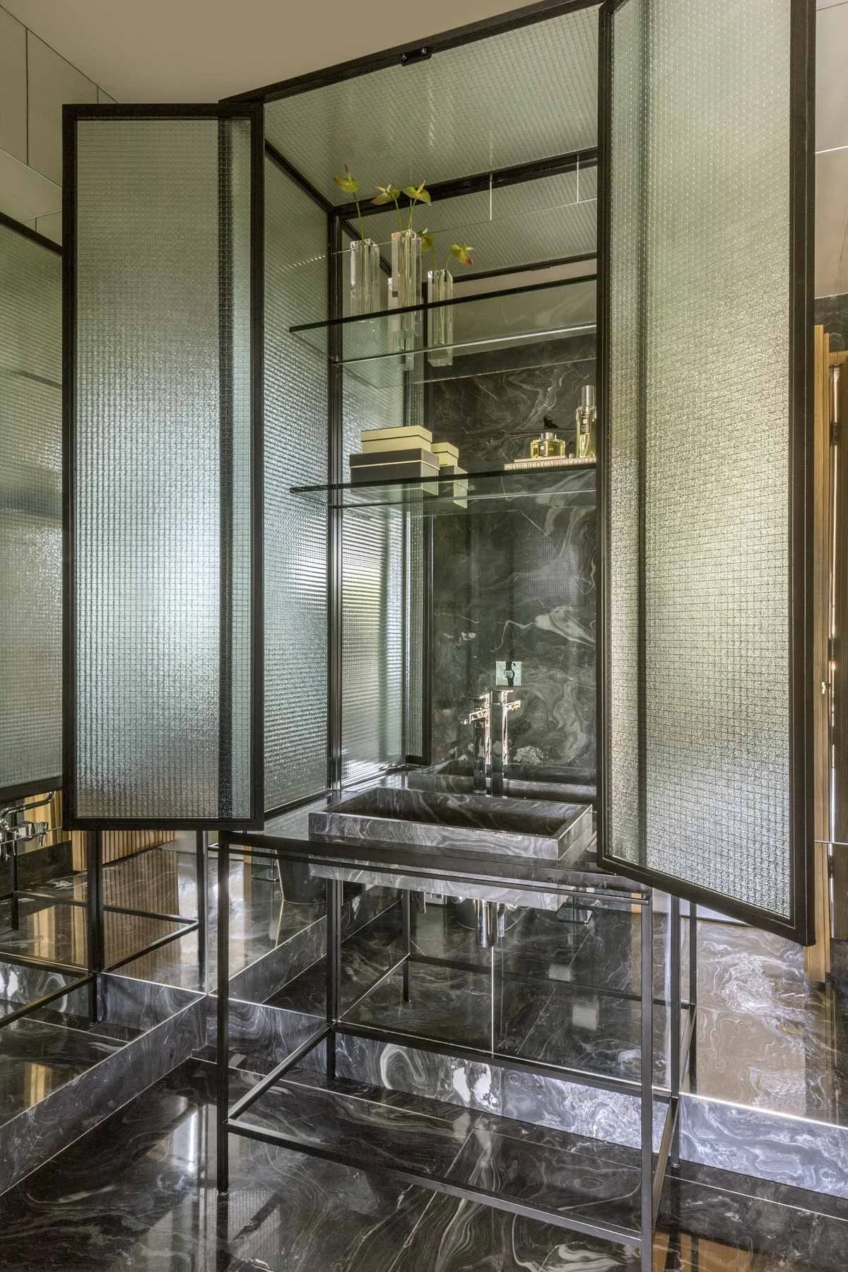 A bathroom vanity features a dark marble basin on a black metal frame, backed by a large mirror and fluted glass cabinet doors.